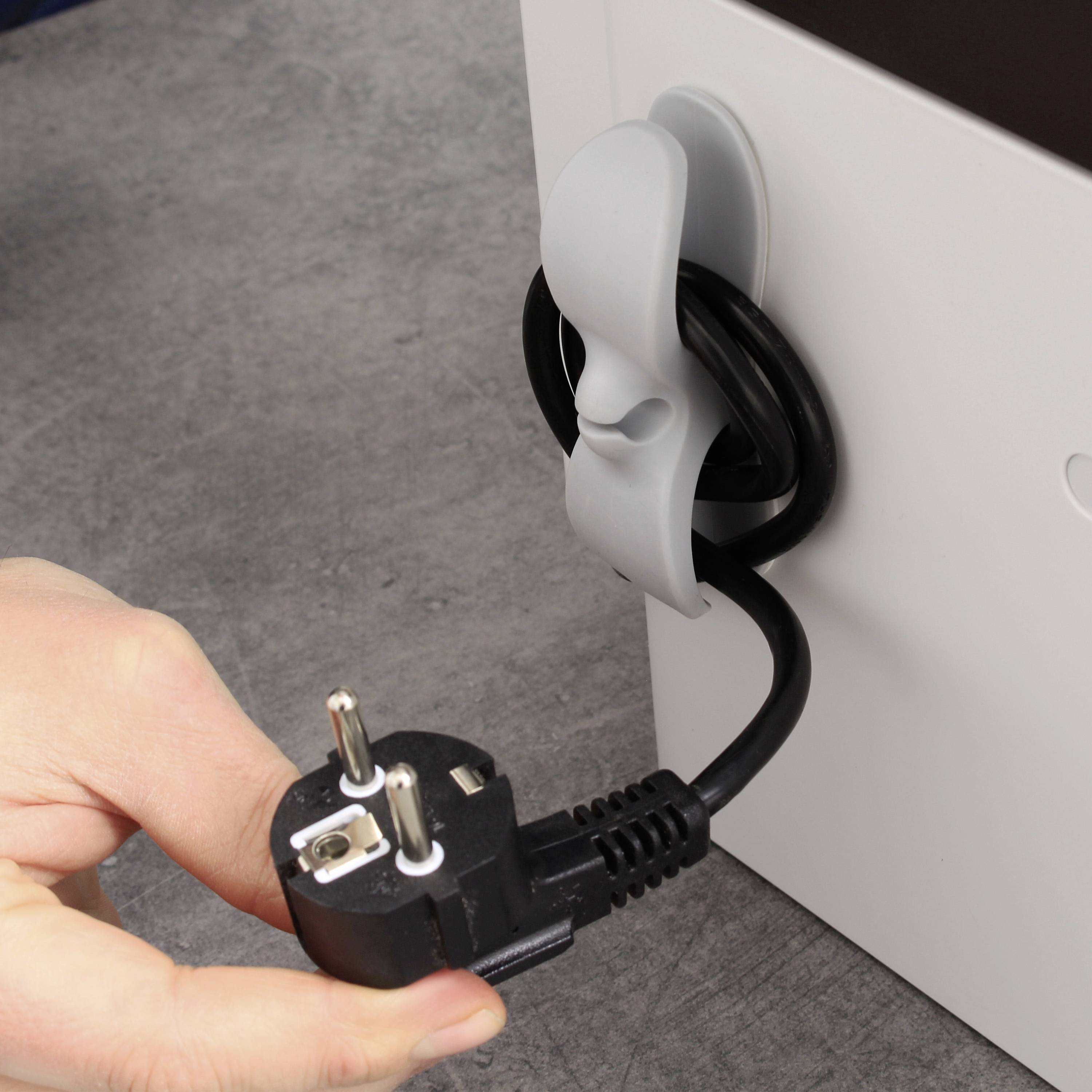 Eine Hand hält einen Stromstecker vor ein Kabel, das ordentlich aufgerollt und mit einem Kabelhalter an einem Möbelstück befestigt ist.
