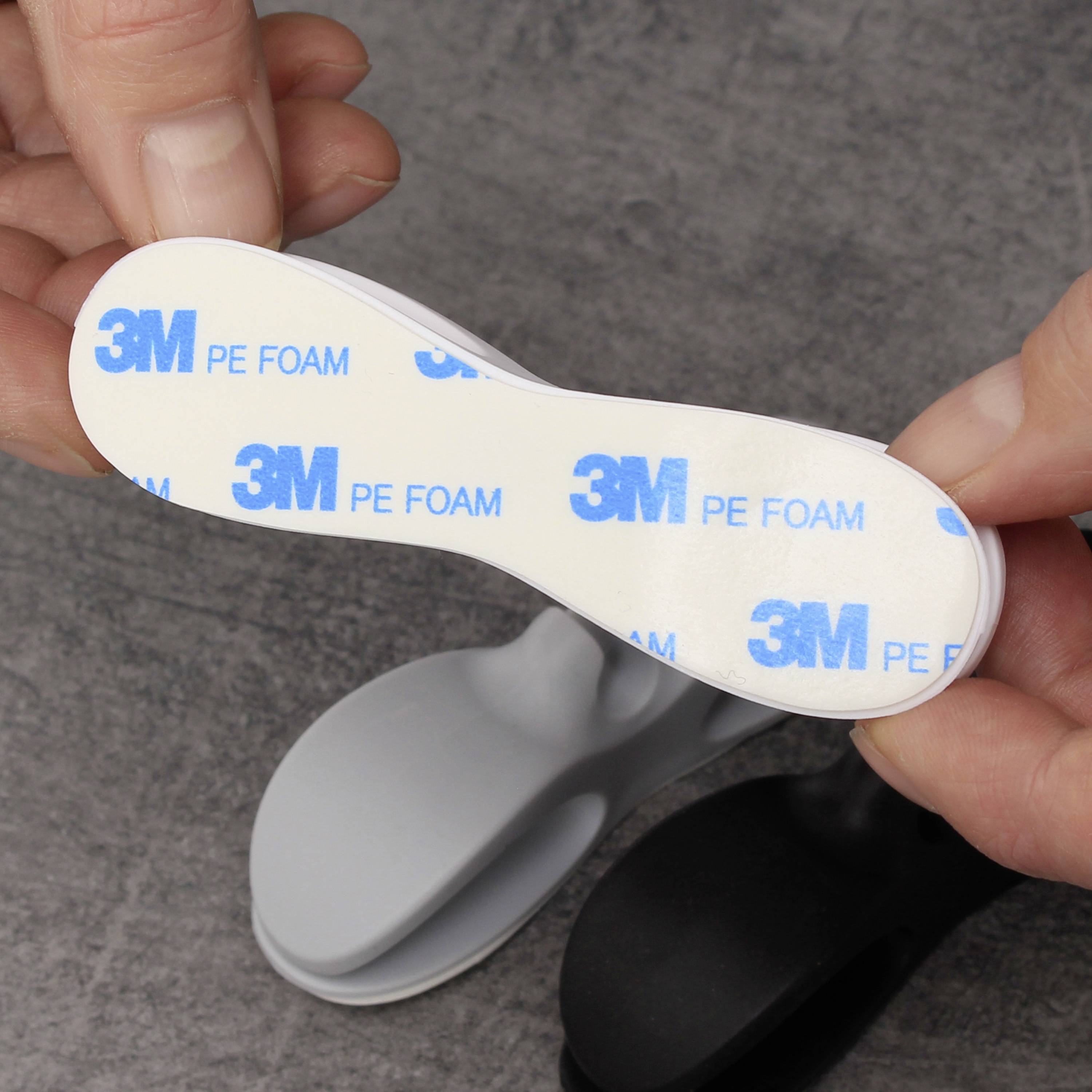 Zwei Hände halten eine kleine, graue Plastikvorrichtung mit 3M PE-Schaumstoff-Aufkleber auf der Unterseite vor grauem Hintergrund.