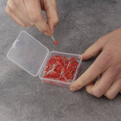 Ein geöffneter Plastikbehälter voller roter LEDs auf einem grauen Hintergrund. Eine Hand hält eine LED, während die andere den Behälter stabilisiert.