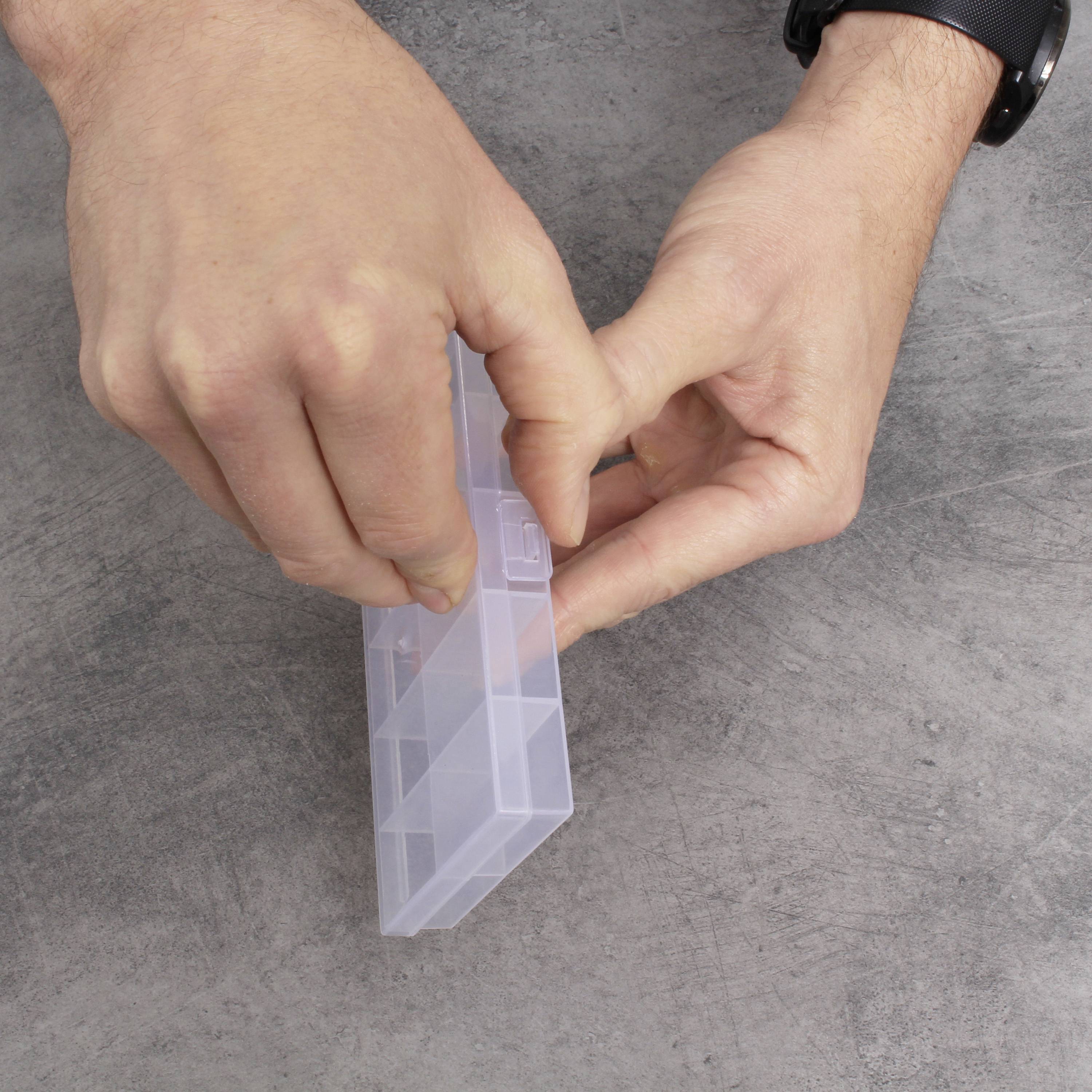 Hände öffnen eine transparente, leere Plastikbox auf einer grauen Oberfläche.