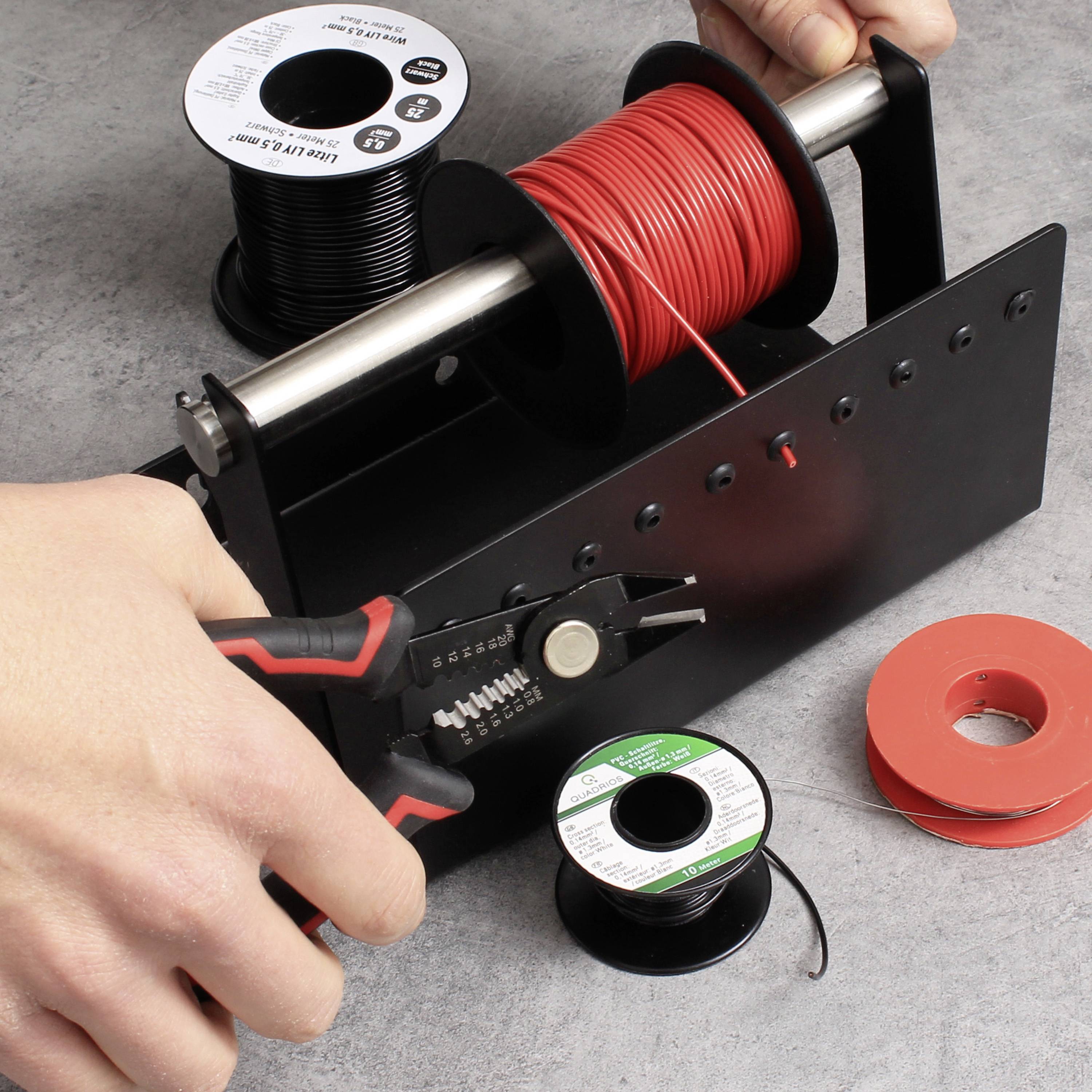 Hand hält Zange, schneidet rotes Kabel von Spule auf Halter, daneben weitere Rollen Isolierband. Arbeitsplatz für Elektriker.