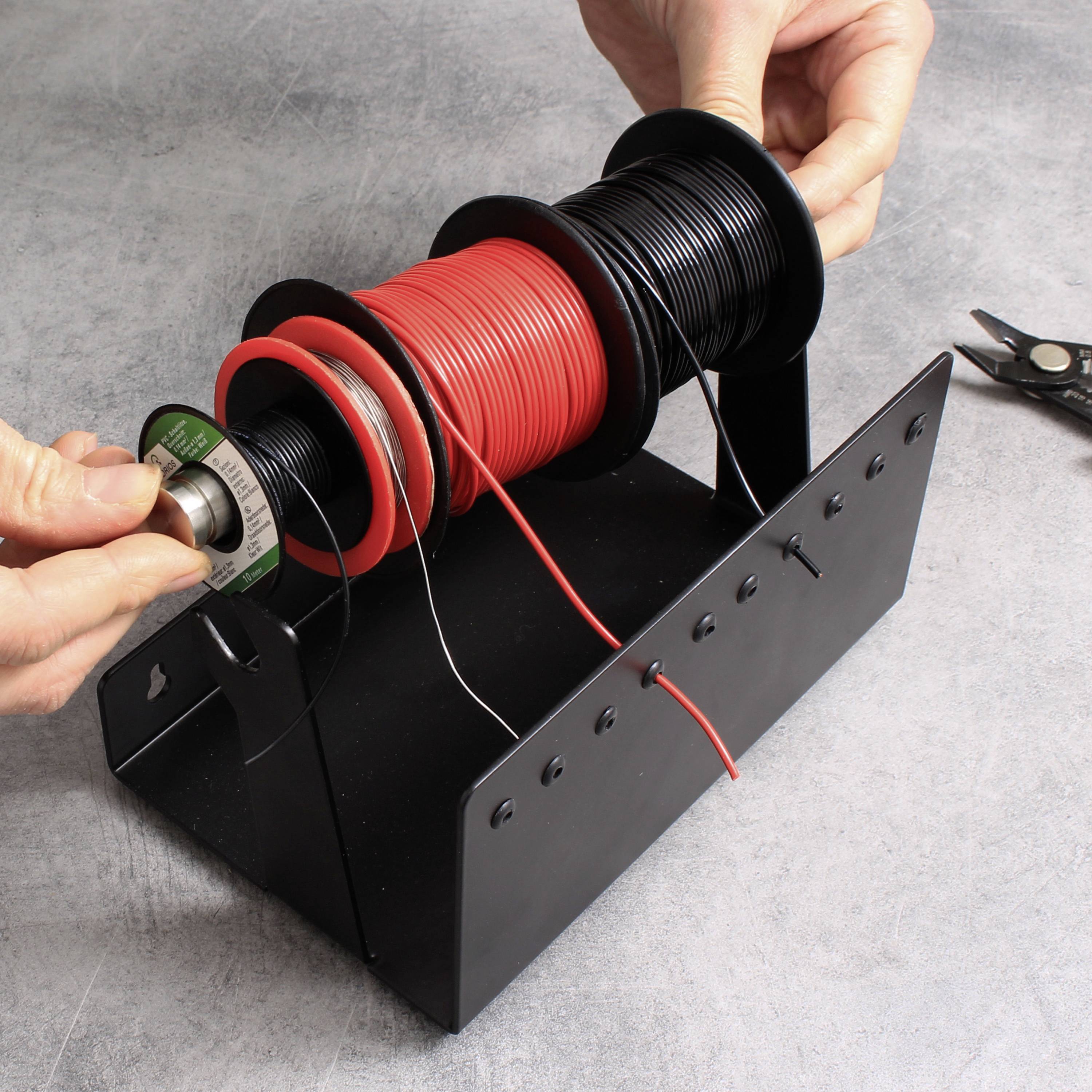 Person zieht Draht von einer Rolle auf einem Tisch ab. Neben der Rolle liegt eine Drahtschere. Die Rollen sind in einem Halter.
