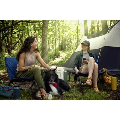 Zwei Personen sitzen entspannt auf Campingstühlen im Wald neben einem Zelt, sie lachen und haben einen schwarzen Hund bei sich.