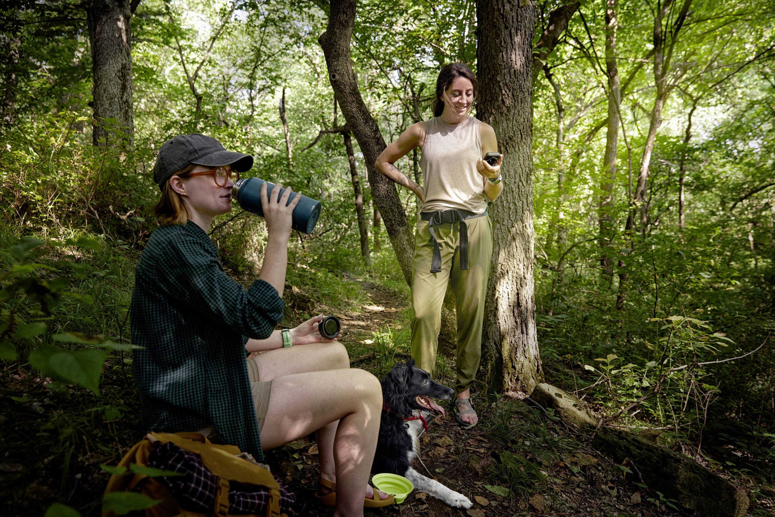 Zwei Personen und ein Hund machen eine Pause im Wald. Eine Person sitzt und trinkt aus einer Flasche, die andere steht mit einem Smartphone.
