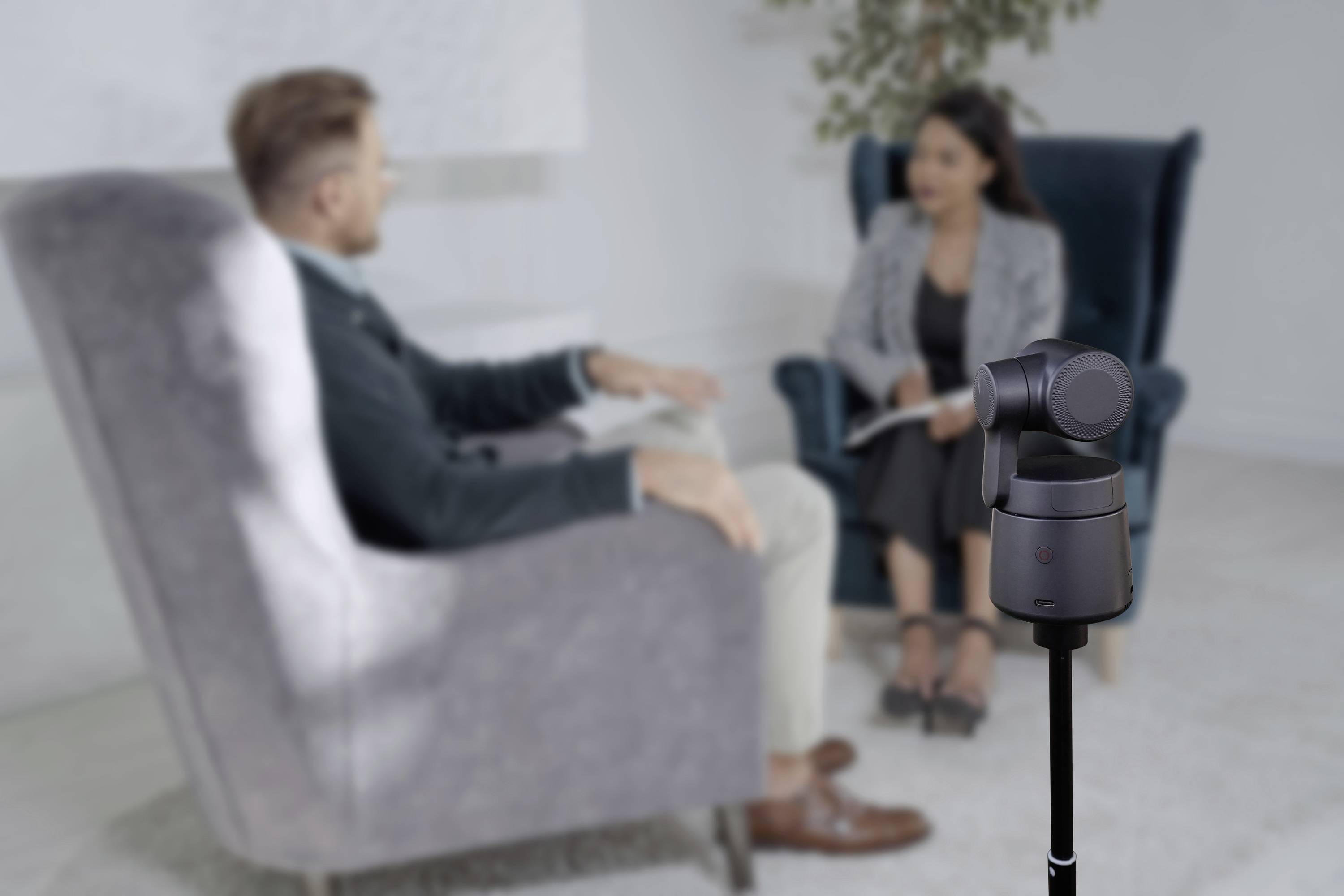 Ein Mann und eine Frau sitzen einander gegenüber in Sesseln. Im Vordergrund steht eine Kamera auf einem Stativ.