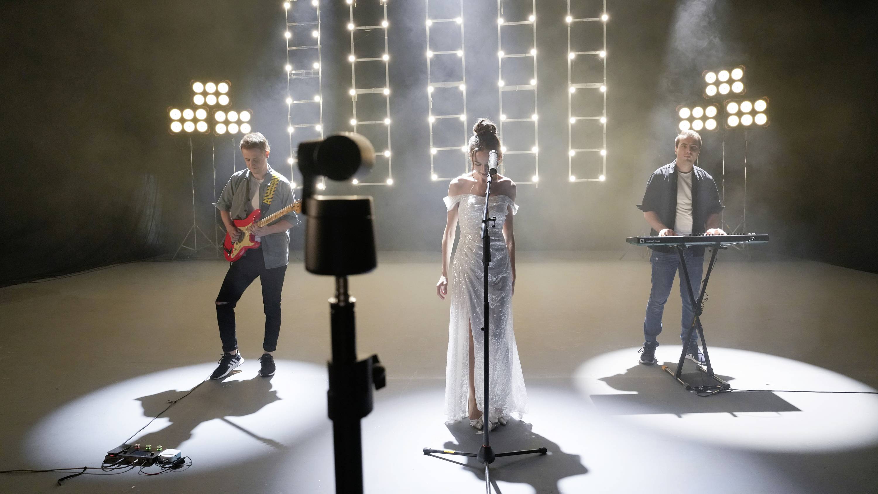 Eine Band performt auf einer Bühne: Sängerin in der Mitte, links ein Gitarrist, rechts ein Keyboarder. Helle Bühnenbeleuchtung im Hintergrund.