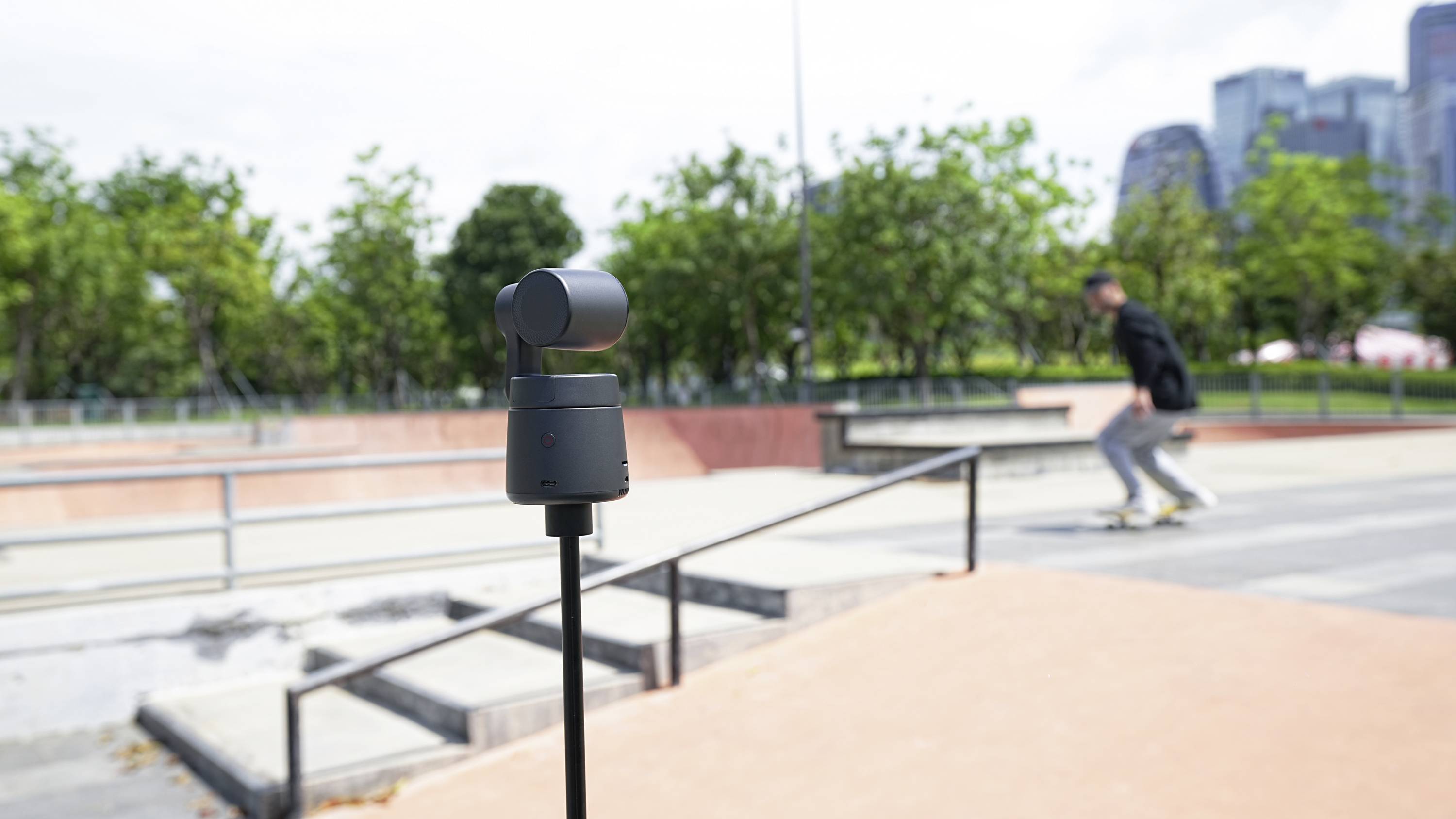 Eine Kamera auf einem Stativ filmt einen Skatepark, während ein Skateboarder in Bewegung im Hintergrund zu sehen ist.