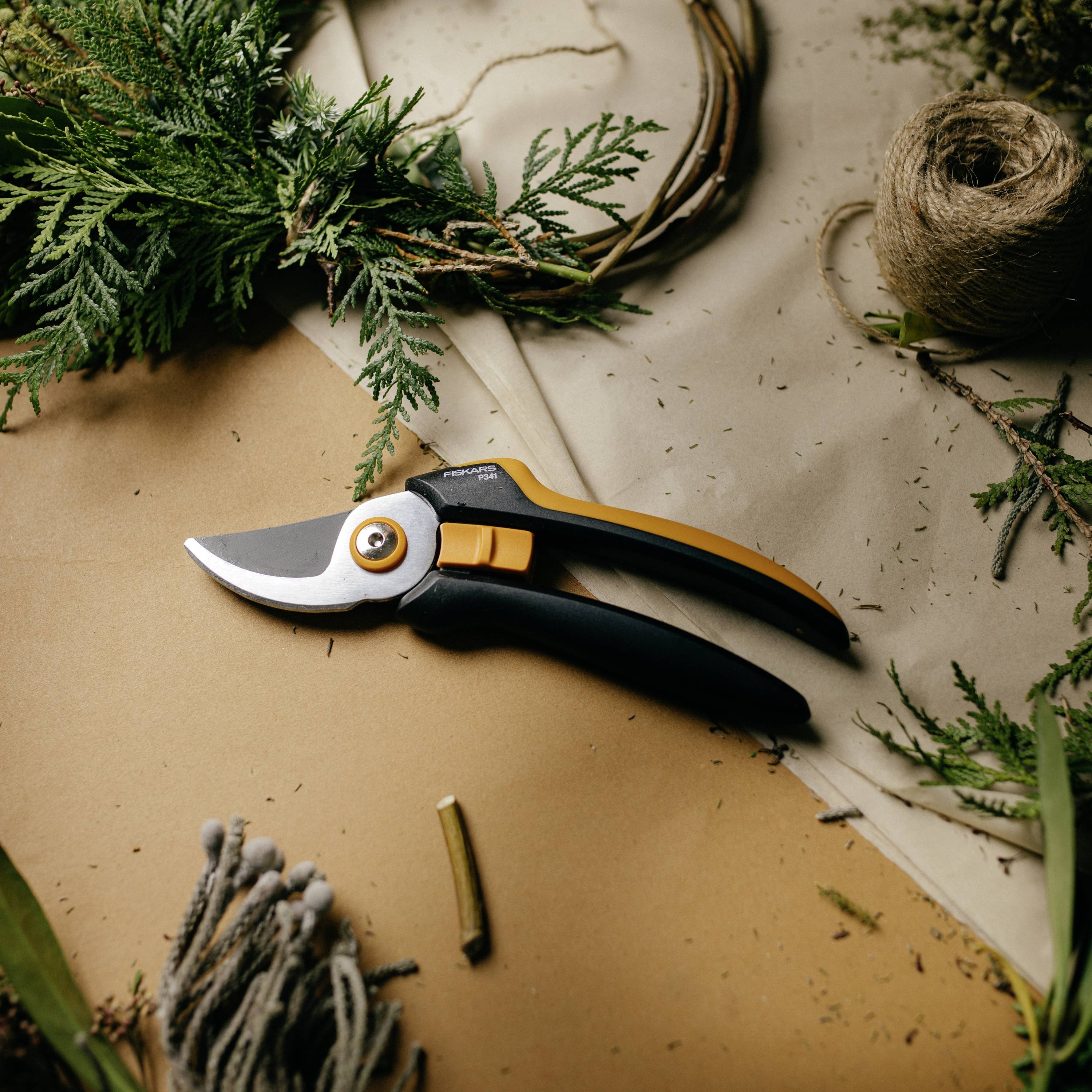 Eine Gartenschere liegt auf einem Tisch, umgeben von Tannenzweigen und Bast, was auf kreatives Basteln oder Floristik hindeutet.