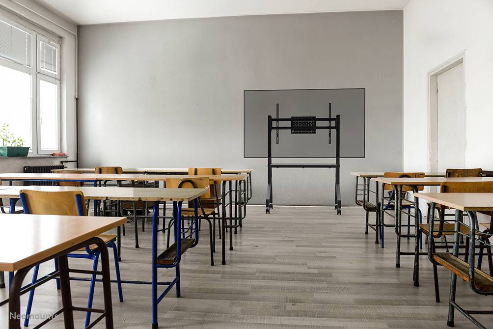 Ein leerer Klassenzimmer mit Stühlen und Tischen. Im Hintergrund steht ein Bildschirmhalter an der grauen Wand. Fenster auf der linken Seite.