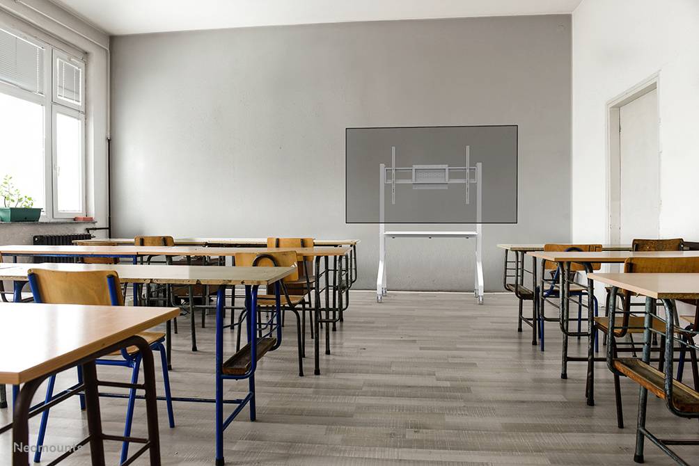 Ein leeres Klassenzimmer mit Tischen und Stühlen, Blick auf eine Wand mit digitalem Whiteboard, Tageslicht von einem großen Fenster.