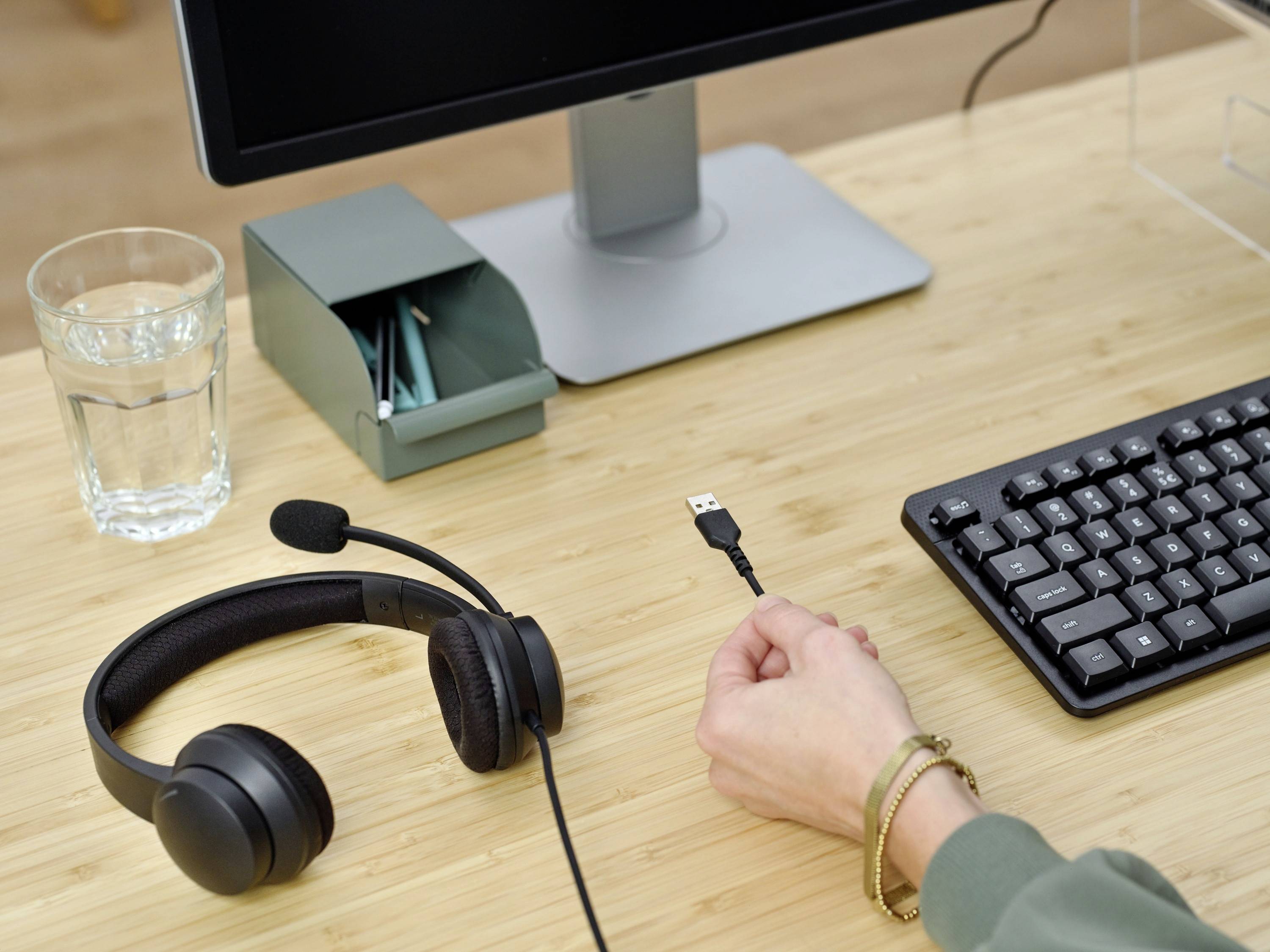 Eine Hand hält ein USB-Stecker neben einer Tastatur und einem Headset auf einem Schreibtisch mit Glas Wasser und Stiften.