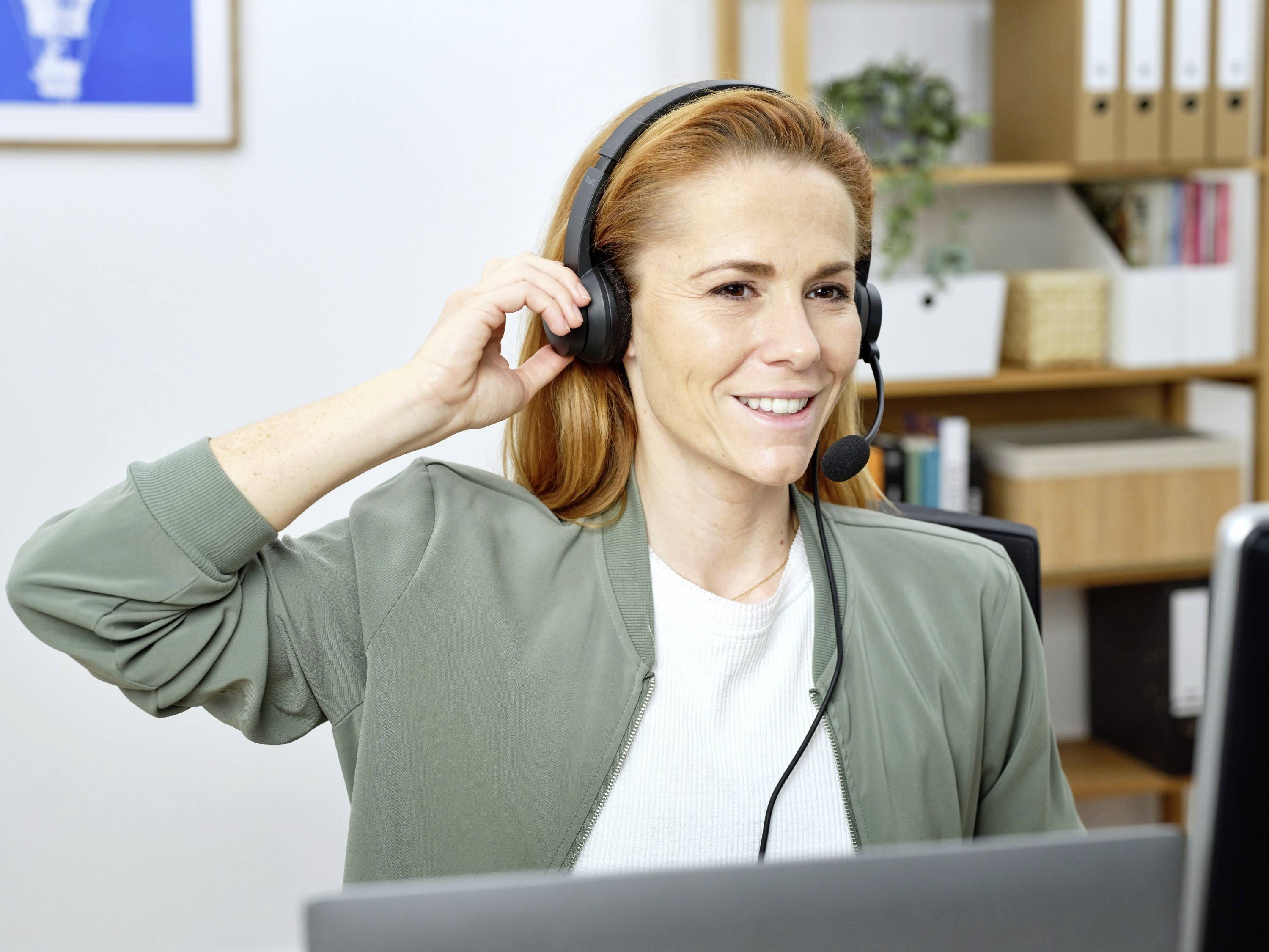 Eine Frau mit roten Haaren trägt ein Headset und lächelt, während sie an einem Schreibtisch sitzt und auf einen Bildschirm schaut.
