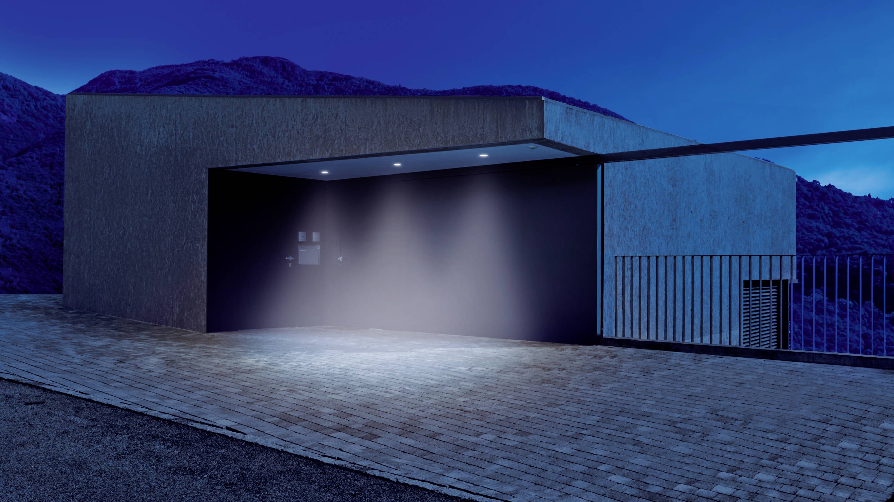 Ein modernes, minimalistisch gestaltetes Gebäude in der Dämmerung mit Betonwänden und einer beleuchteten Einfahrt.