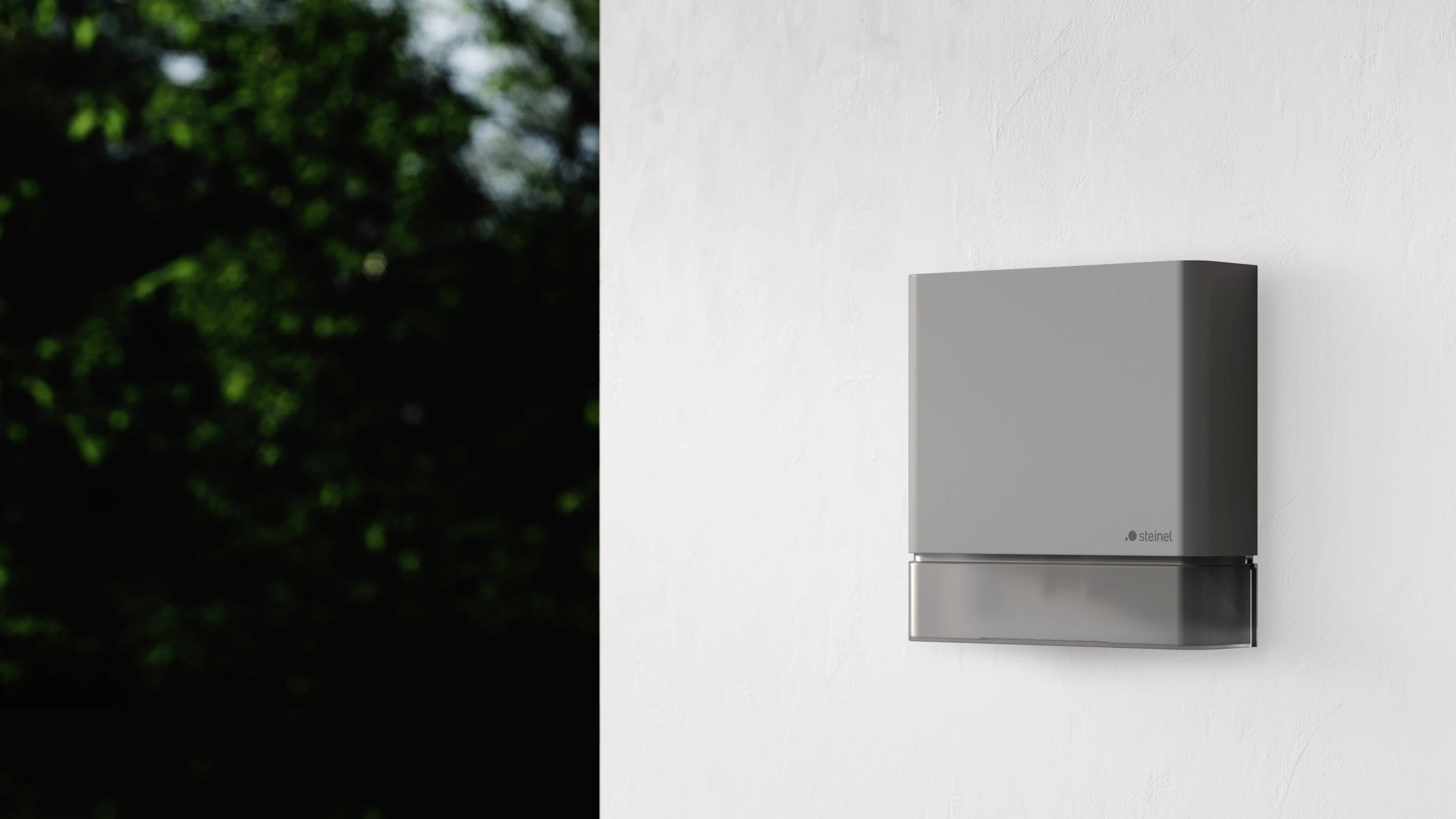 Moderne, wandmontierte Klimaanlage in grauem Gehäuse an weißer Wand, grüne Pflanzen im Hintergrund sichtbar.