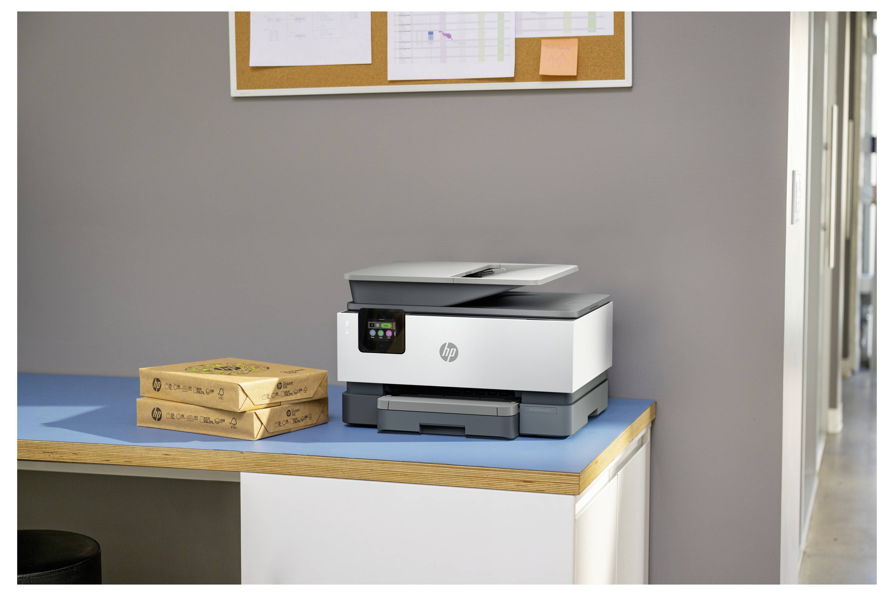 HP Multifunktionsdrucker auf einem Schreibtisch neben zwei gestapelten Kartonpaketen in einer Büroumgebung.