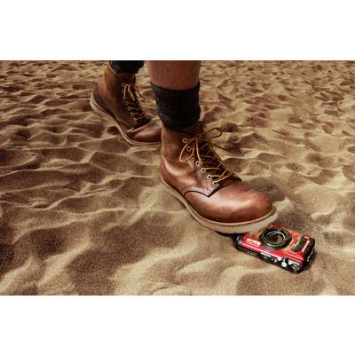 Ein großer Stiefel tritt auf ein rotes Spielzeugauto im Sand, das teilweise unter dem Schuh eingequetscht ist.