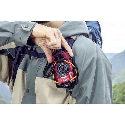 Ein Mann in Outdoor-Kleidung trägt eine rote Kamera um den Hals und zeigt mit der Hand eine Geste. Hinter ihm sind Berge sichtbar.