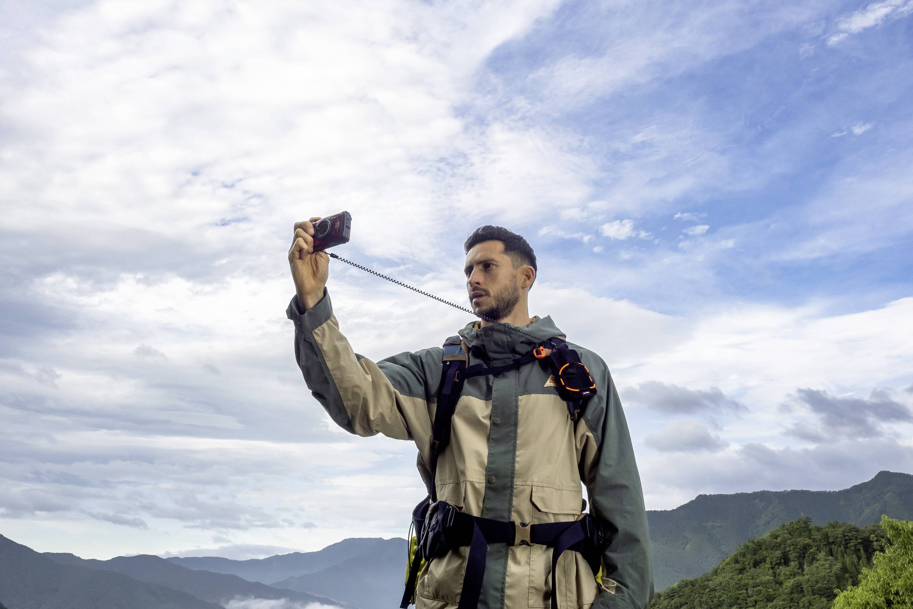 Ein Mann in Outdoor-Kleidung macht ein Selfie mit dem Smartphone vor einer Kulisse aus Bergen und bewölktem Himmel.