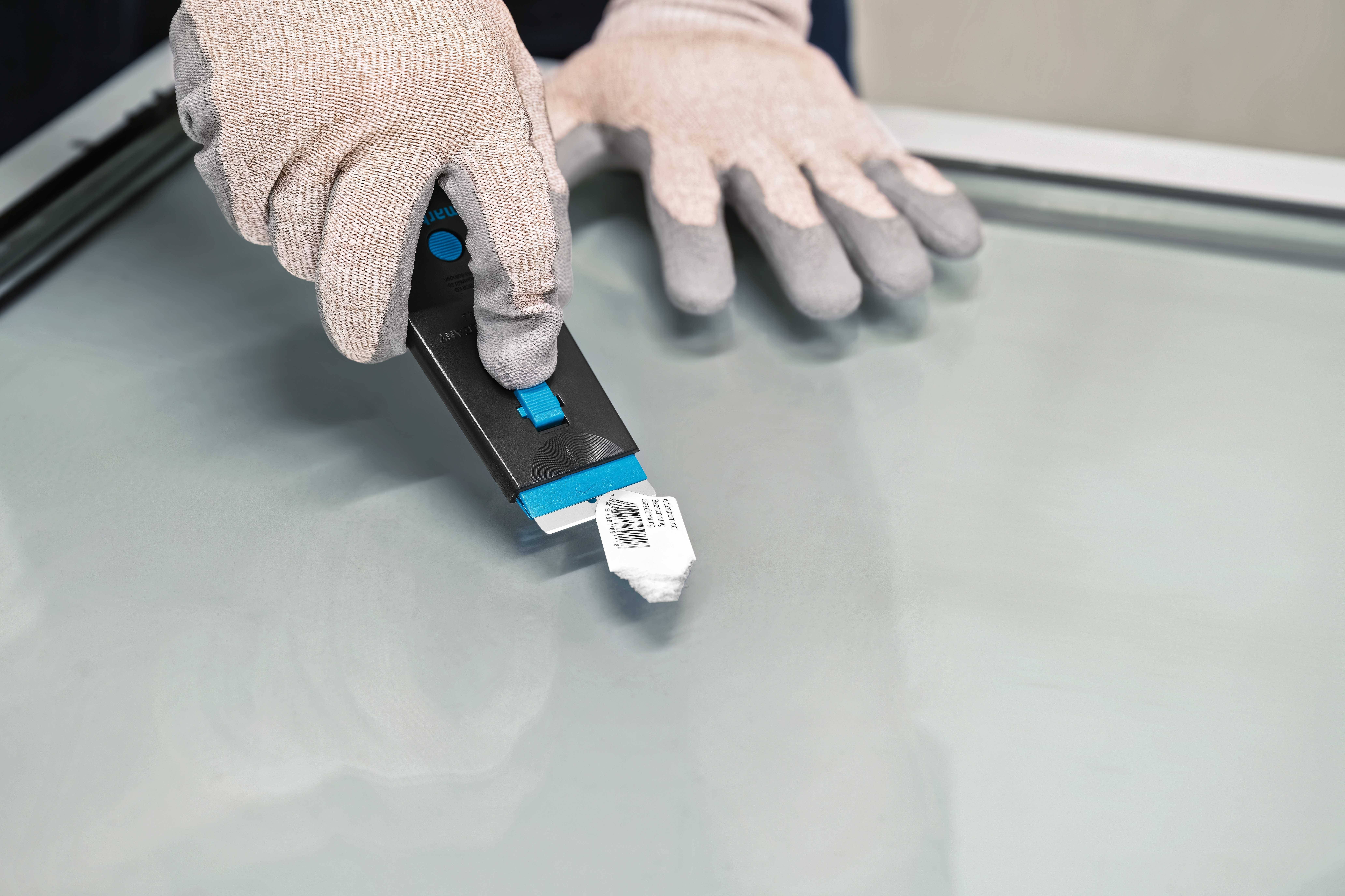 Eine Person entfernt mit einem Handabstreifer vorsichtig einen Aufkleber von einer Glasoberfläche. Sie trägt Sicherheitshandschuhe.