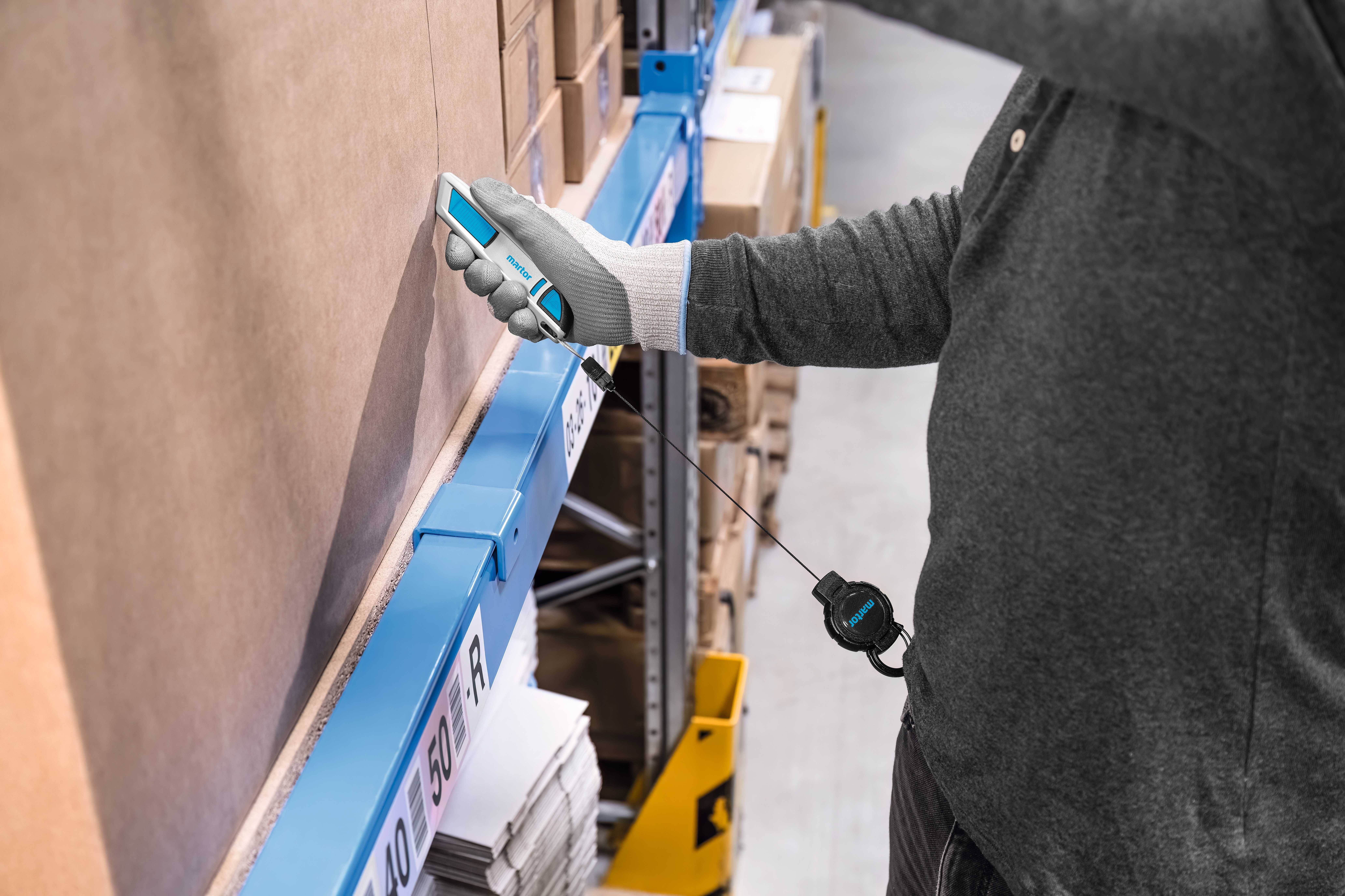 Eine Person im Lager scannt mit einem Handscanner ein Produkt auf einem Regal. Sie trägt Arbeitshandschuhe und ein graues Oberteil.