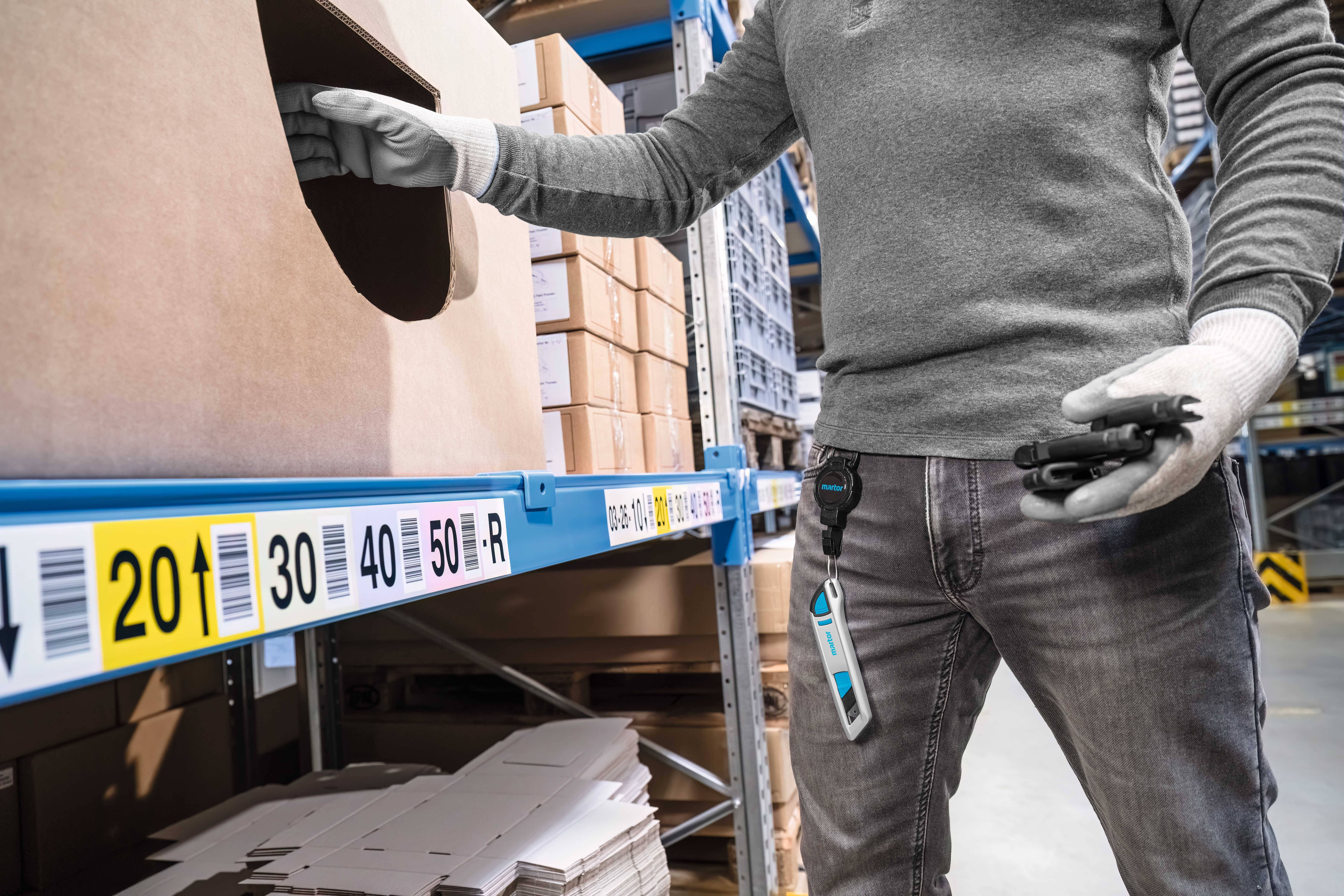Eine Person mit Handschuhen holt einen Gegenstand aus einem Regal im Lager. Regale mit Kartons und Etiketten mit Zahlen sind sichtbar.
