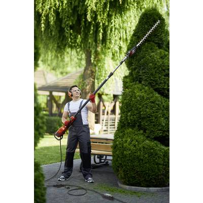 Eine Person schneidet mit einem elektrischen Heckenschneider eine hohe, spitz geformte Hecke in einem Garten.