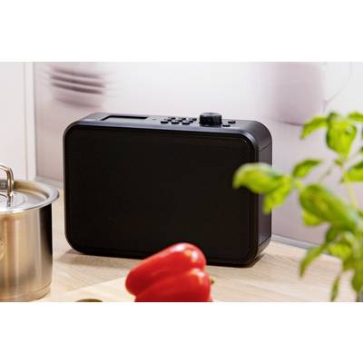 Ein schwarzes Radio steht auf einer Küchenarbeitsfläche neben einem Topf, roten Paprika und Basilikum.