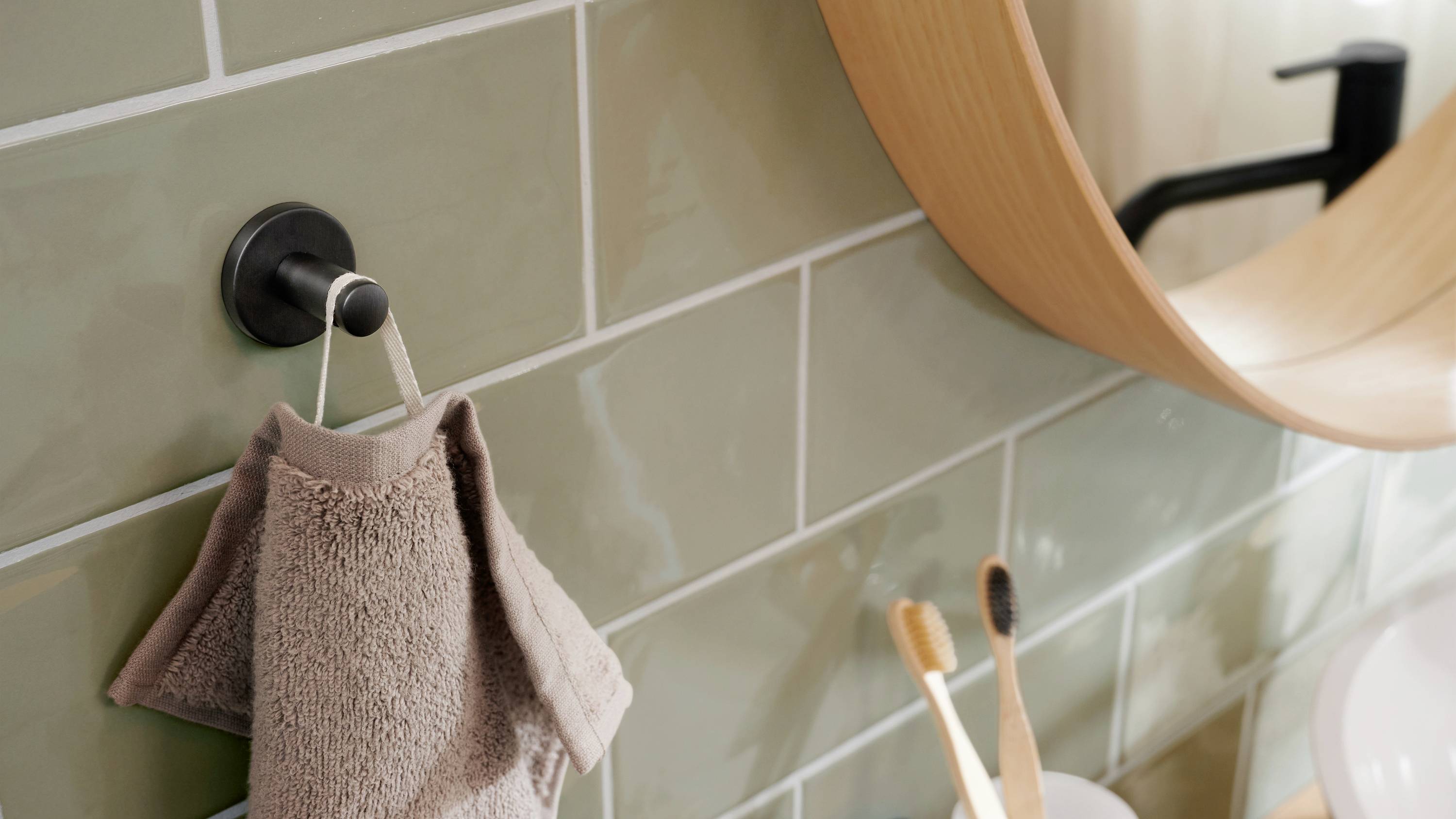 Ein beige Handtuch hängt an einem Haken an der grünen Kachelwand, daneben stehen zwei Zahnbürsten im Becher am Waschbecken.