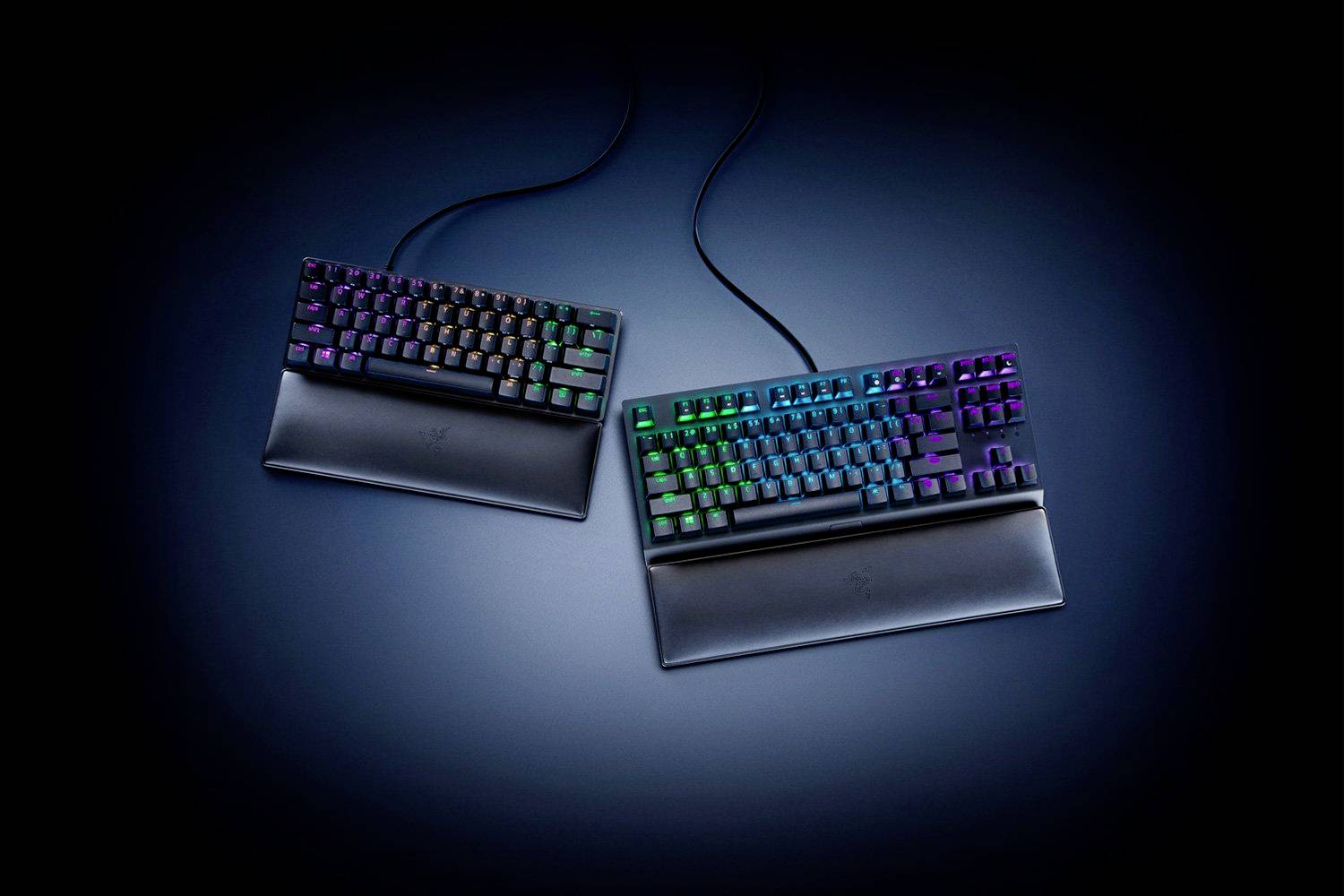 Zwei mechanische Tastaturen mit RGB-Hintergrundbeleuchtung auf dunklem Hintergrund. Beide haben abnehmbare Handballenauflagen und Kabel.