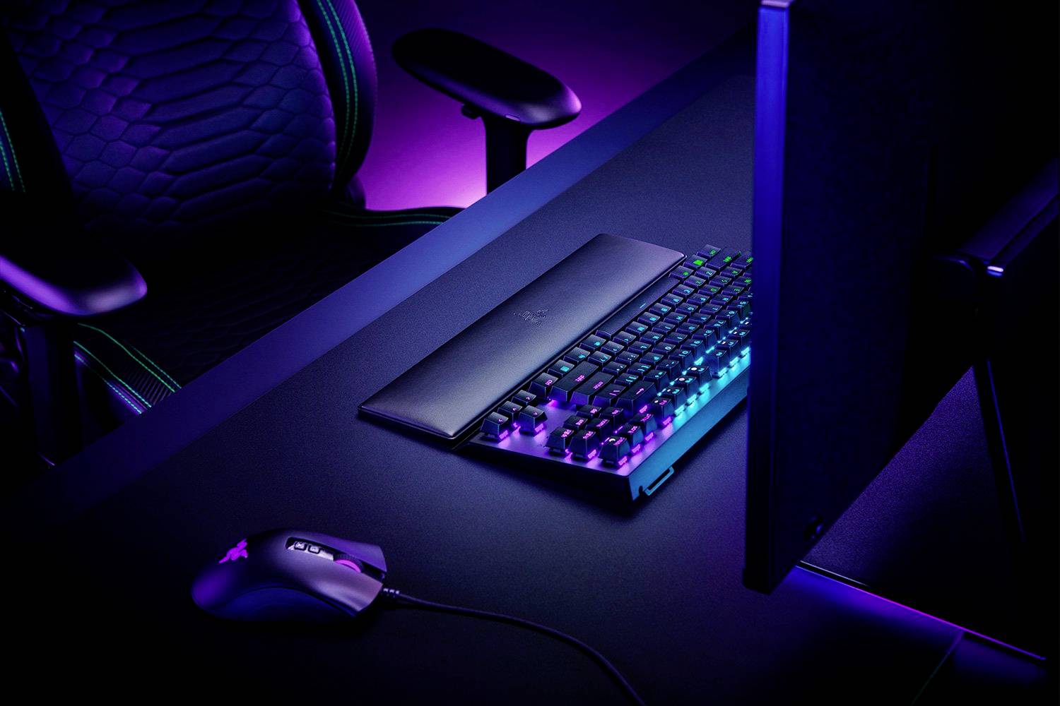Ein Gaming-Setup mit beleuchteter Tastatur, Gaming-Maus und Bildschirm auf einem Schreibtisch, von violettem Licht stimmungsvoll erhellt.