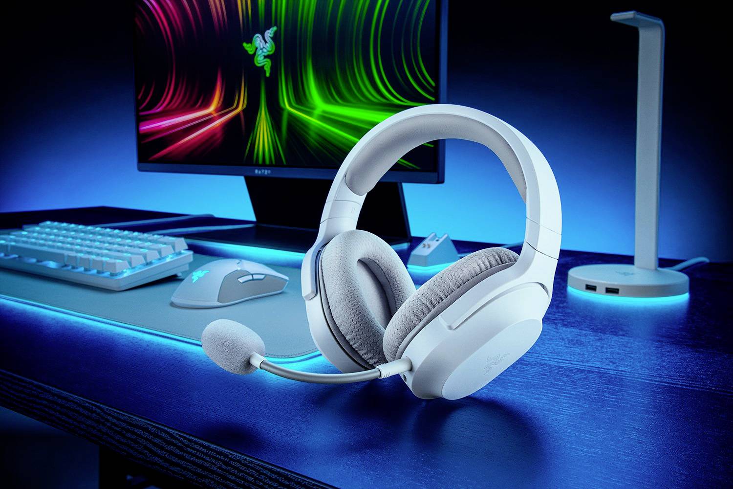 Ein weißes Gaming-Headset liegt auf einem Schreibtisch vor einem Monitor mit leuchtenden, bunten Wellen. Im Hintergrund ist eine Tastatur.