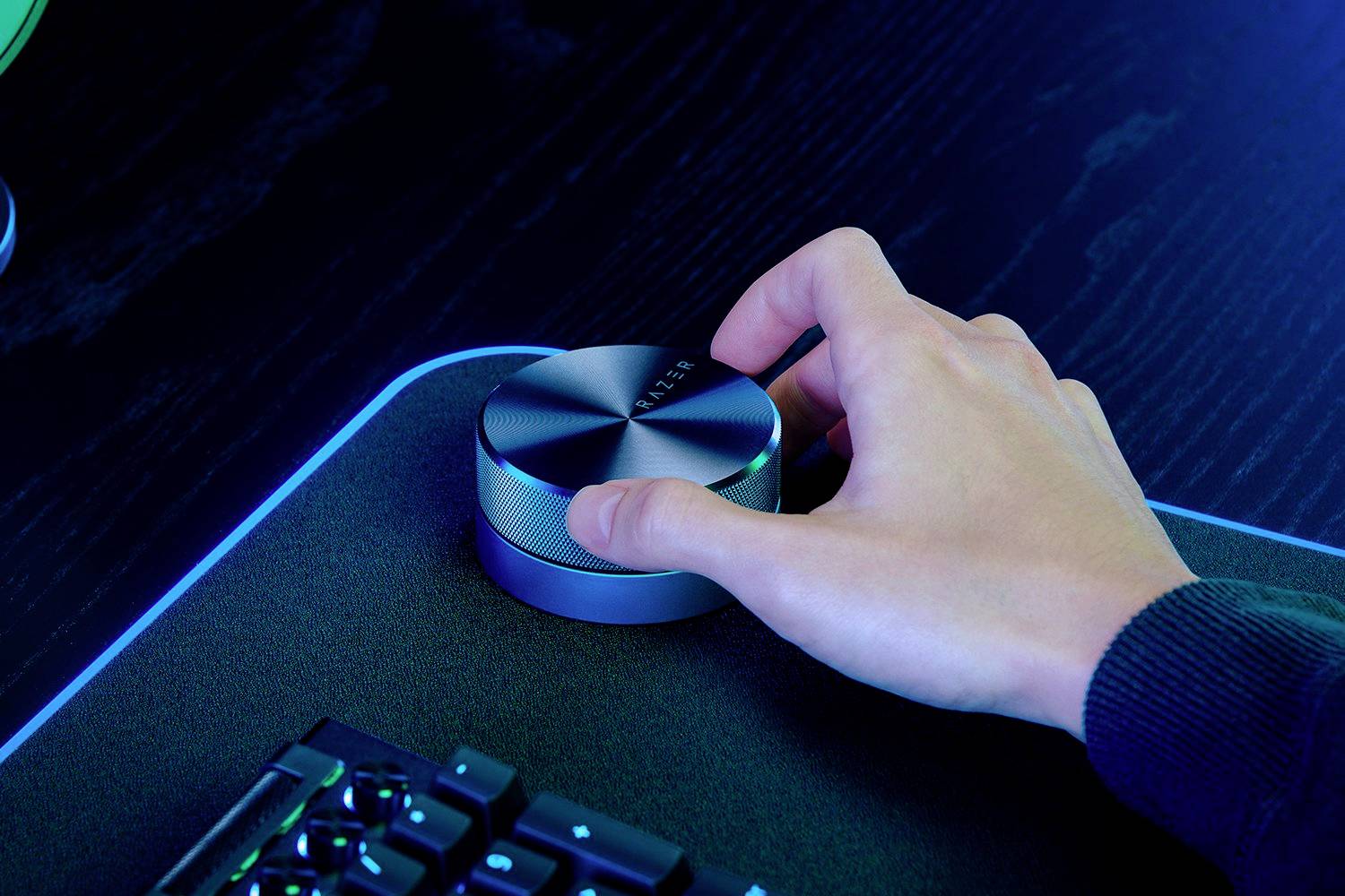 Hand dreht an einem silbernen, runden Lautstärkeregler auf einem Schreibtisch neben einer Tastatur auf einem beleuchteten Pad.