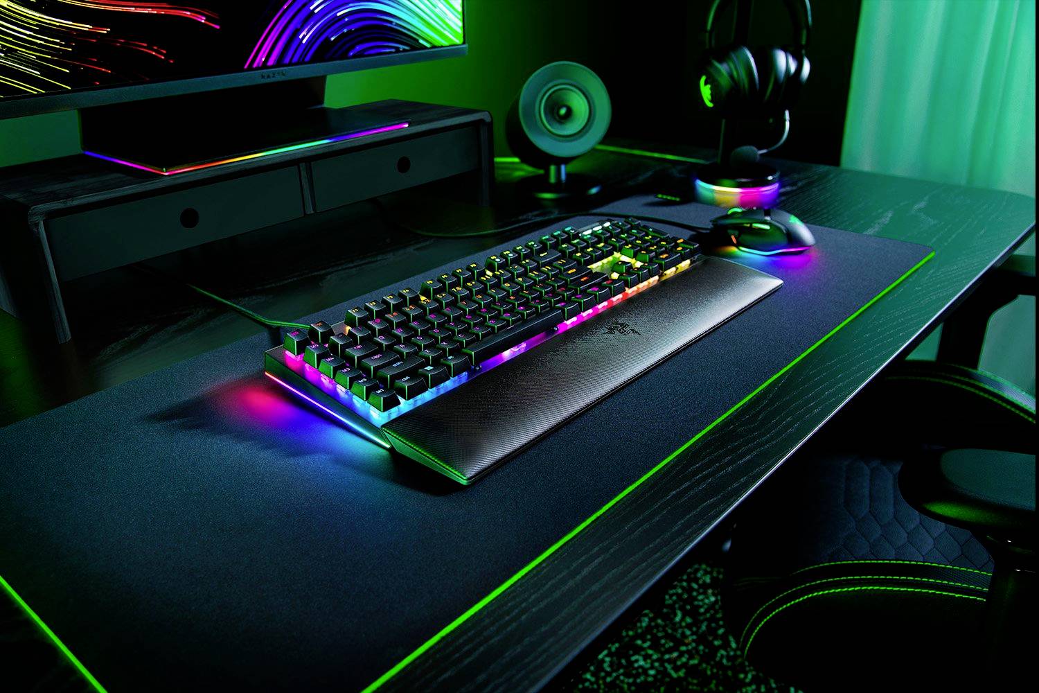 Ein Gaming-Schreibtisch mit bunter Tastaturbeleuchtung, Monitor, Lautsprecher und Kopfhörer. Die Beleuchtung ist in Regenbogenfarben.