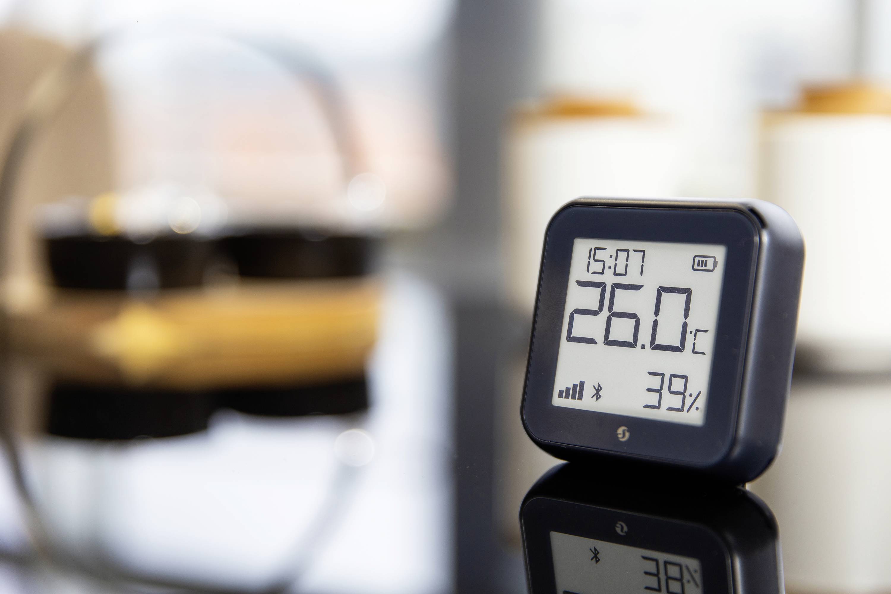 Ein digitaler Thermo-Hygrometer zeigt 26,0°C und 39% Luftfeuchtigkeit an, Zeit 15:07. Im Hintergrund unscharf Haushaltsgegenstände.