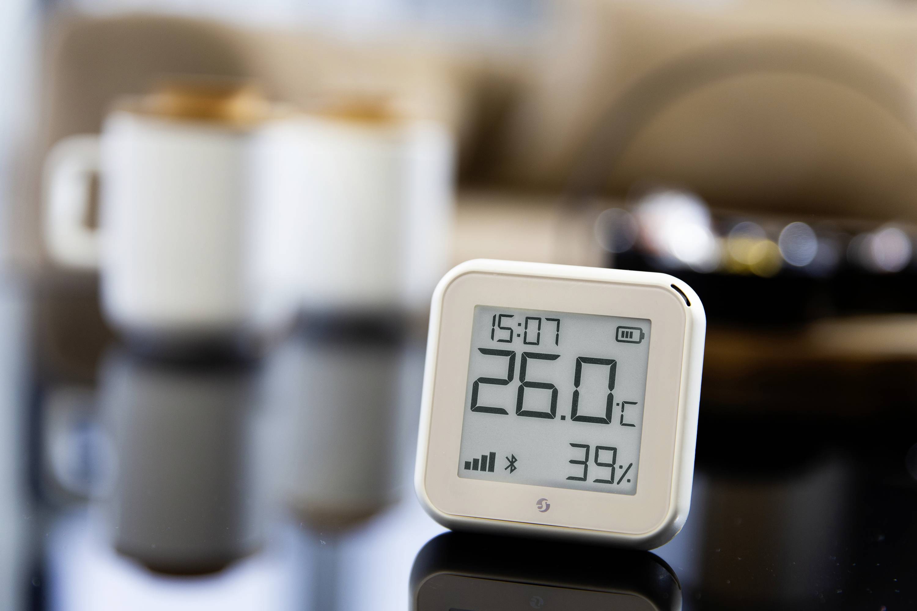 Digitales Thermo-Hygrometer zeigt 26,0°C und 39% Luftfeuchtigkeit an, im Hintergrund verschwommene Tassen.