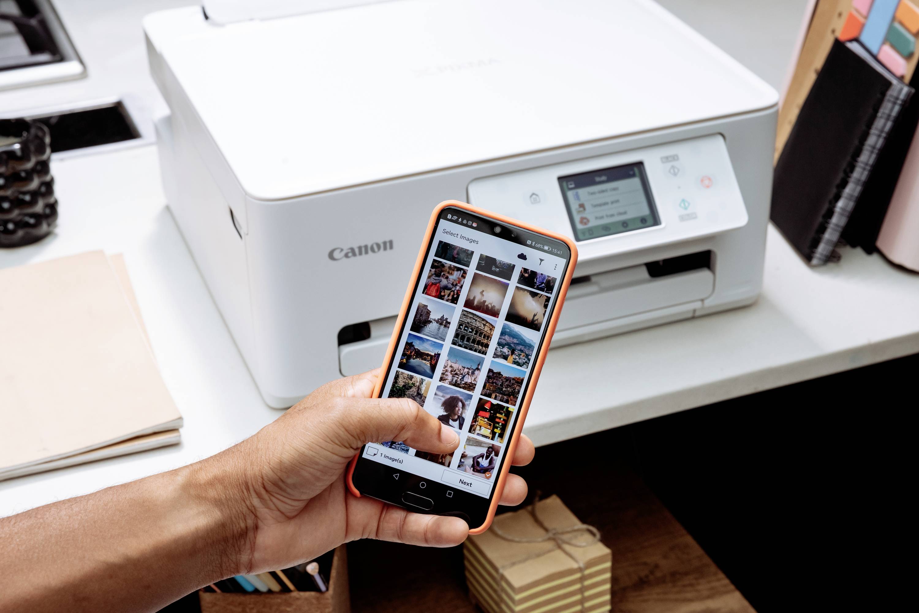 Person hält ein Smartphone vor einem Drucker. Auf dem Handybildschirm sind Fotos zu sehen. Der Drucker steht auf einem Tisch.