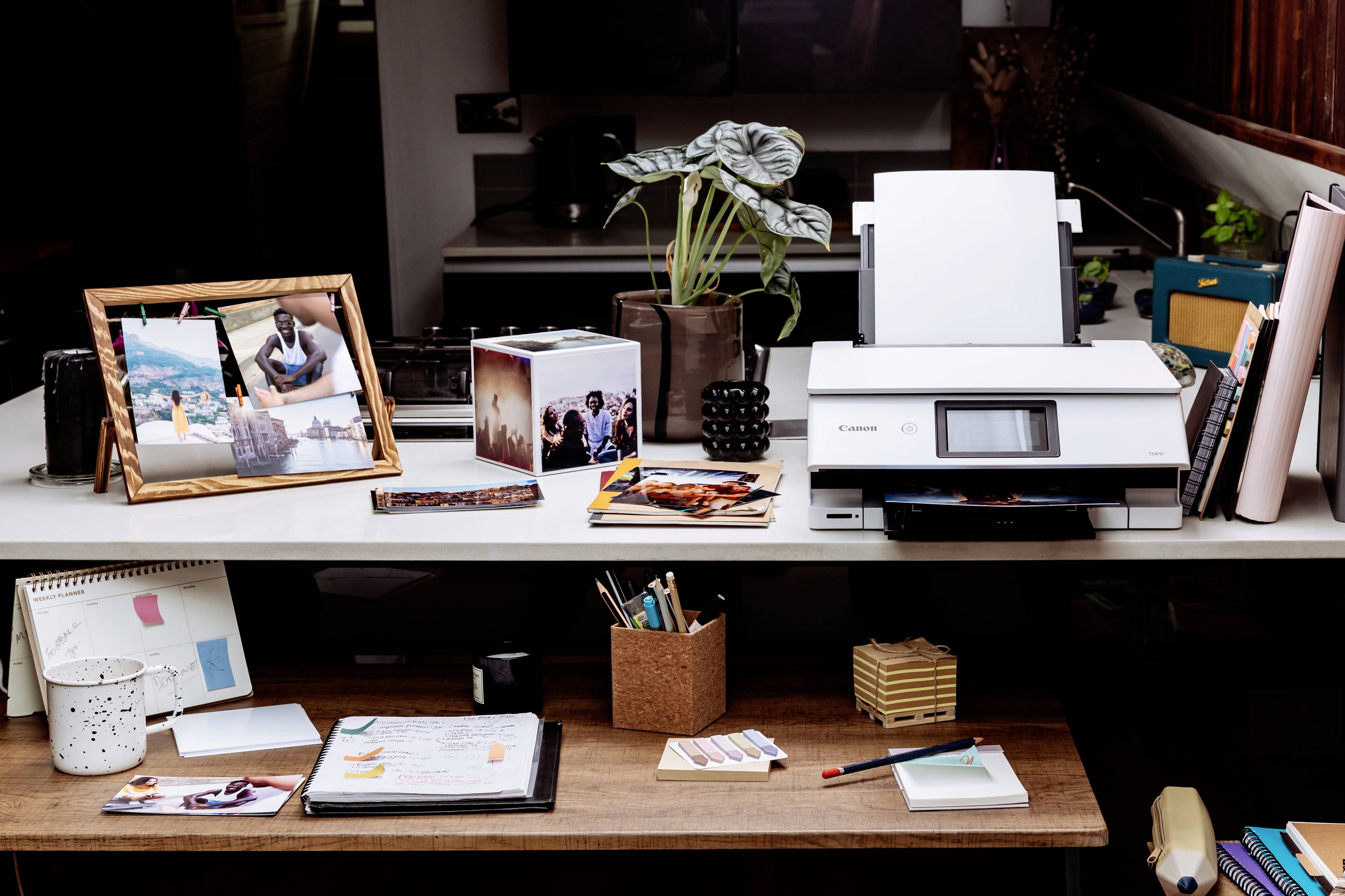 Drucker auf einem Schreibtisch mit Fotos, Stiften und Notizbüchern, umgeben von Pflanzen und Büromaterialien.