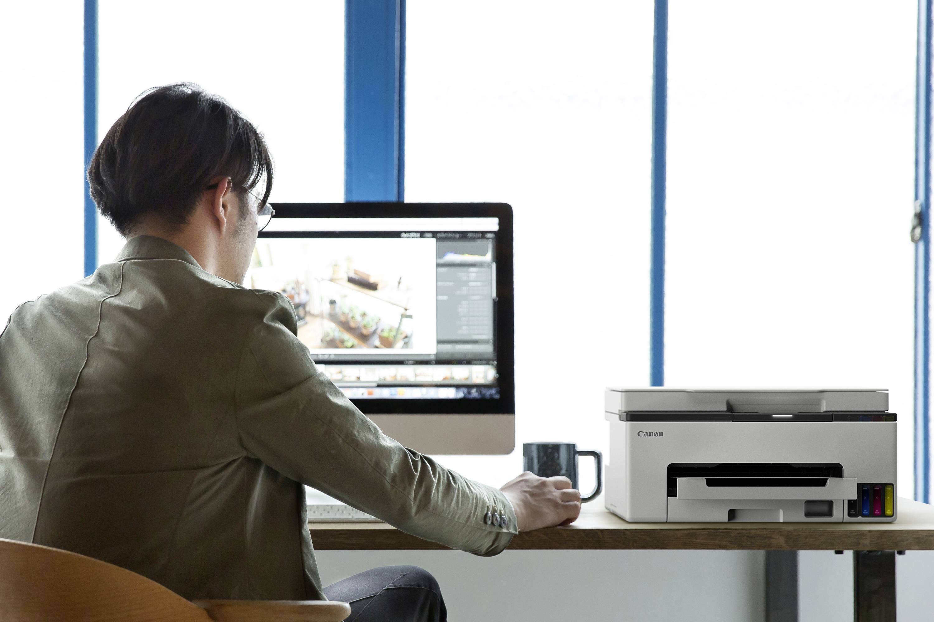 Person sitzt an einem Schreibtisch und arbeitet an einem Computer, eine Tasse neben dem Monitor. Ein Drucker steht daneben.