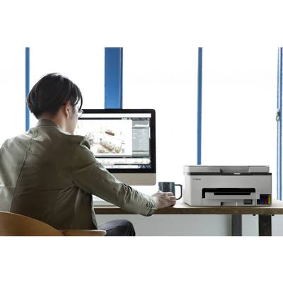 Person sitzt an einem Schreibtisch und arbeitet an einem Computer, eine Tasse neben dem Monitor. Ein Drucker steht daneben.