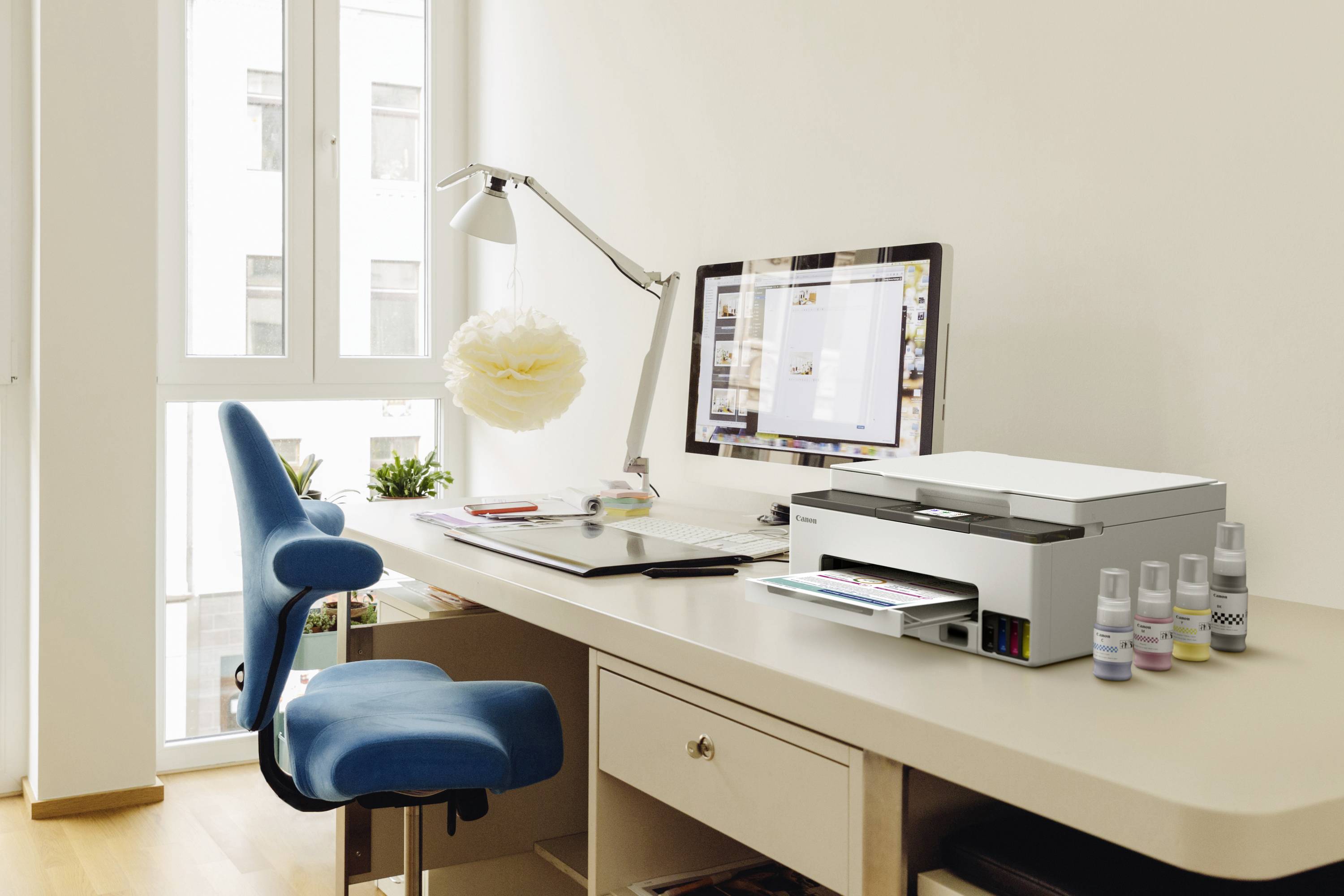 Arbeitsplatz mit einem Computer, Drucker und blauer Bürostuhl auf einem hellen Holztisch, umgeben von Fenstern mit Tageslicht.