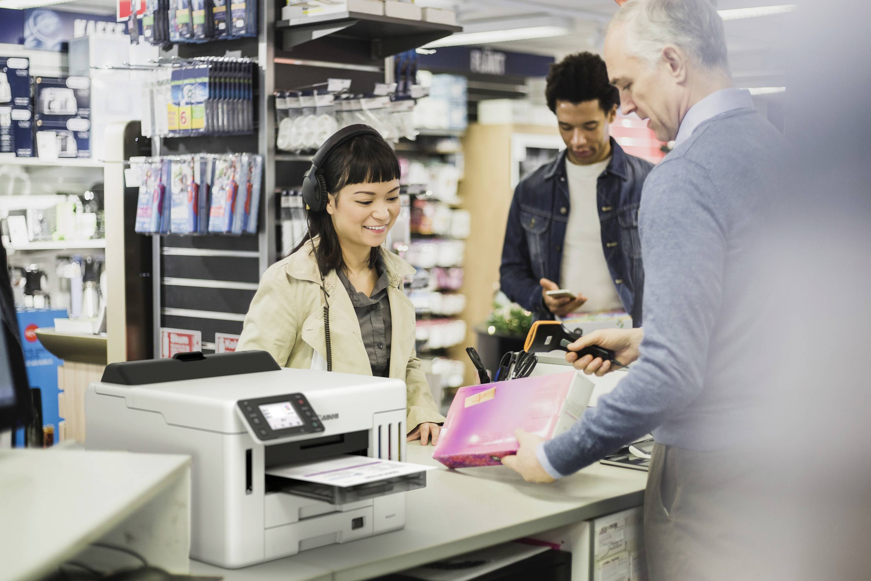 Ein Mann scannt eine Patrone an der Kasse eines Elektronikladens. Verkäuferin lächelt, Kunde im Hintergrund schaut auf sein Handy.