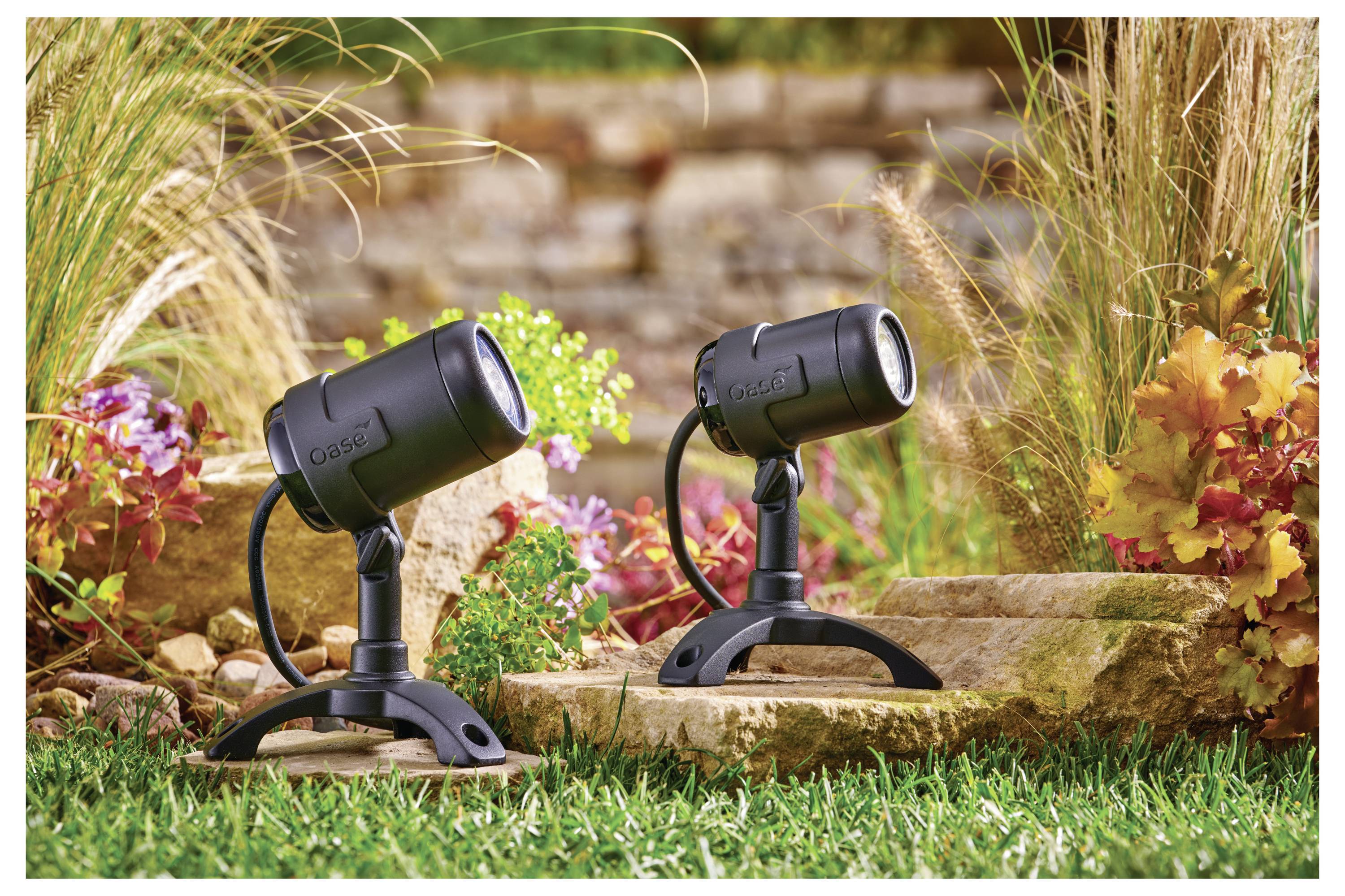 Zwei schwarze Außenstrahler auf einem grünen Rasen mit Herbstpflanzen und einer Steinmauer im Hintergrund, die Gartenbeleuchtung veranschaulichen.