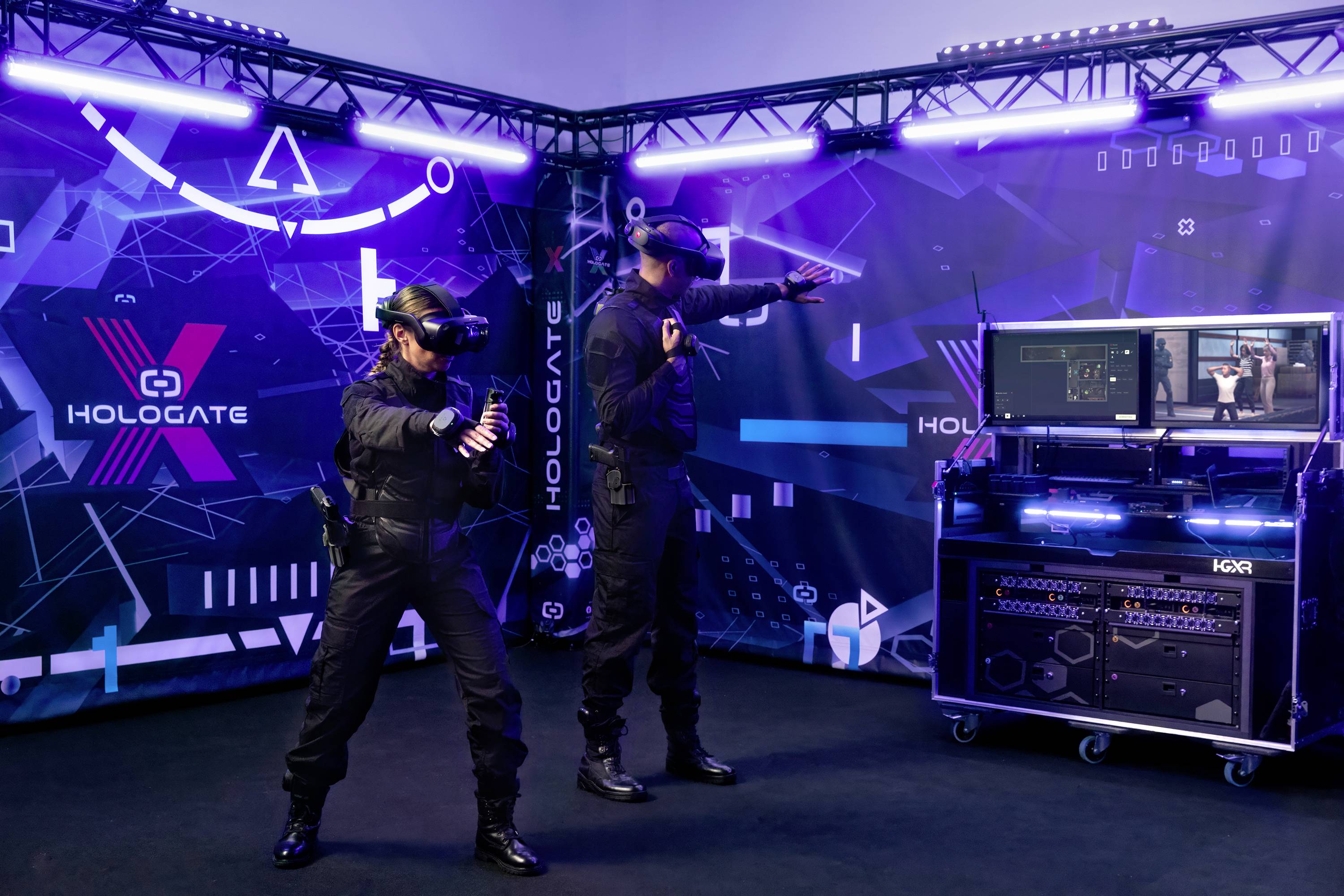 Zwei Personen in Virtual-Reality-Brillen interagieren in einem futuristisch gestalteten Raum mit Hologate-Logo und elektronischer Ausrüstung.