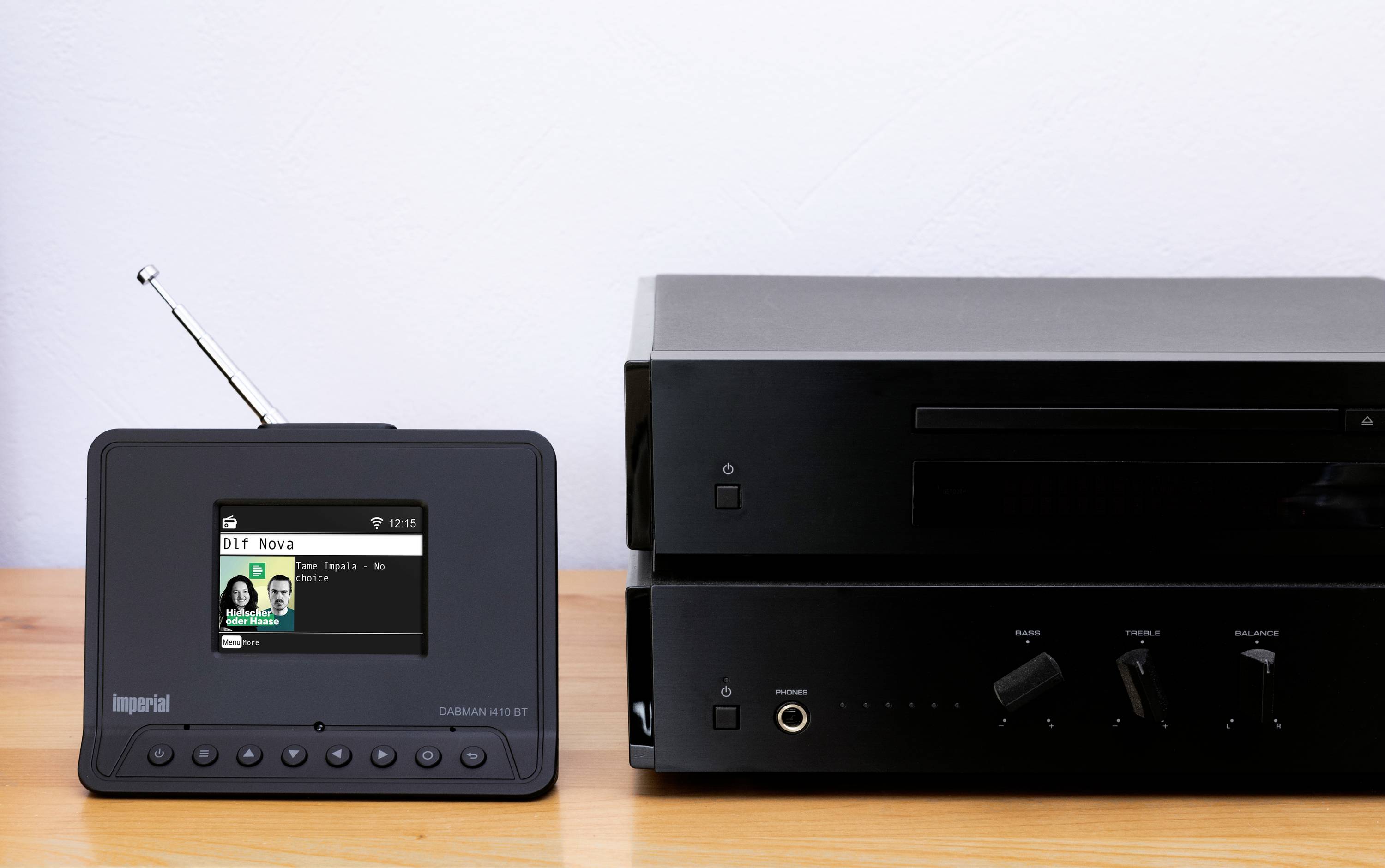 Kompakte schwarze Digitalradio-Anlage neben einem Verstärker auf einem Holzregal. Die Geräte haben Knöpfe und eine Antenne.