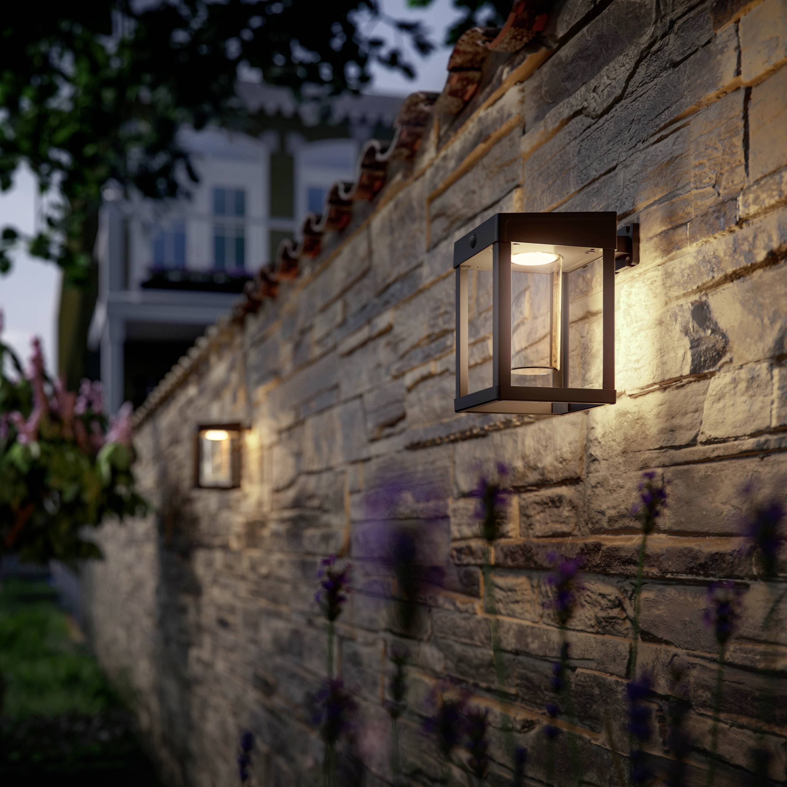 Zwei leuchtende Wandlampen an einer Steinmauer beleuchten einen Gehweg bei Dämmerung. Im Hintergrund sind unscharfe Gebäude sichtbar.