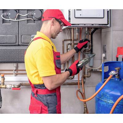 Ein Handwerker in gelber und roter Arbeitskleidung überprüft mit einem Messgerät eine Gasleitung, umgeben von Rohren und einem blauen Gastank.