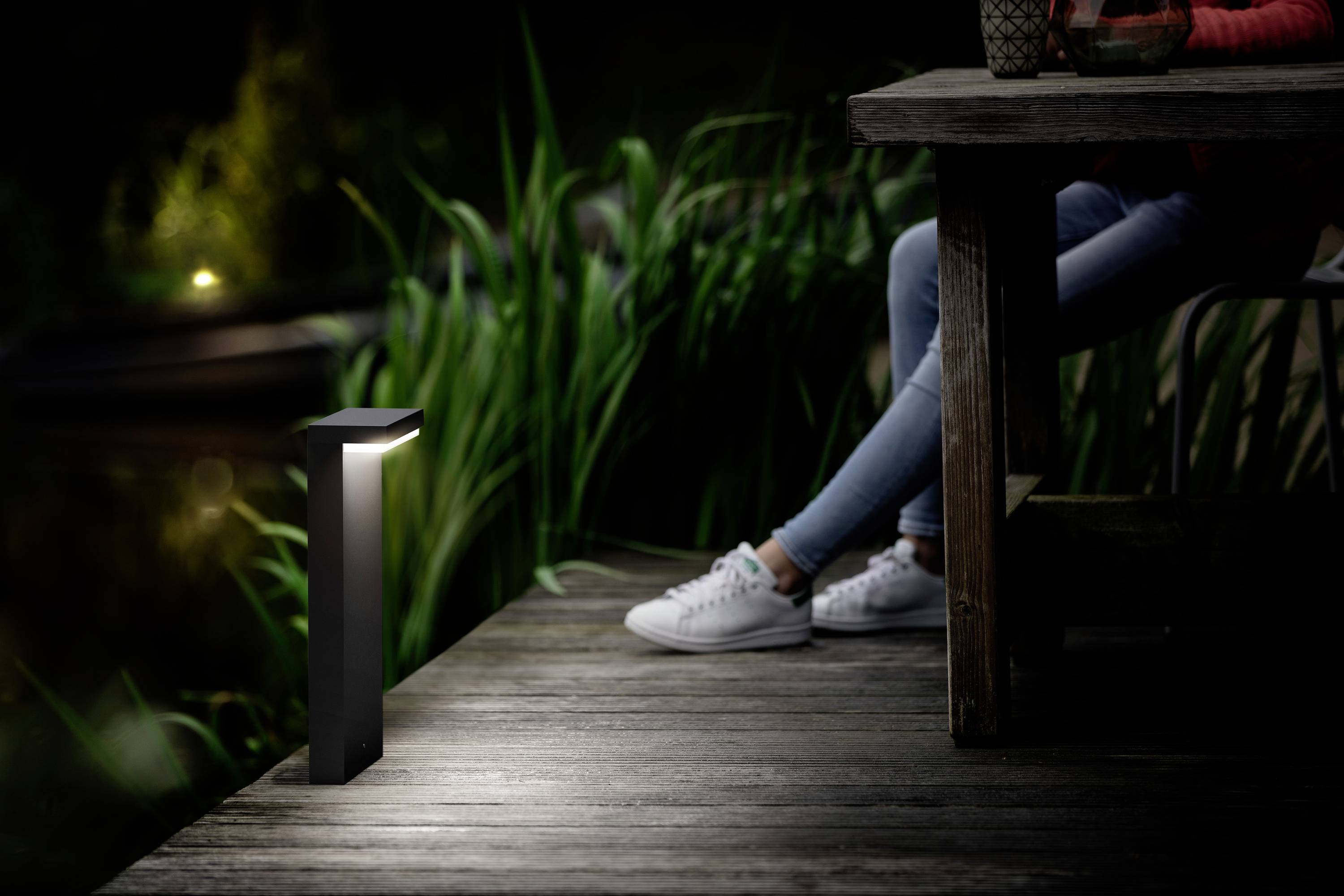 Eine Person sitzt nachts an einem Holztisch auf einer Terrasse. Eine moderne Außenleuchte beleuchtet den Boden neben dem Tisch.