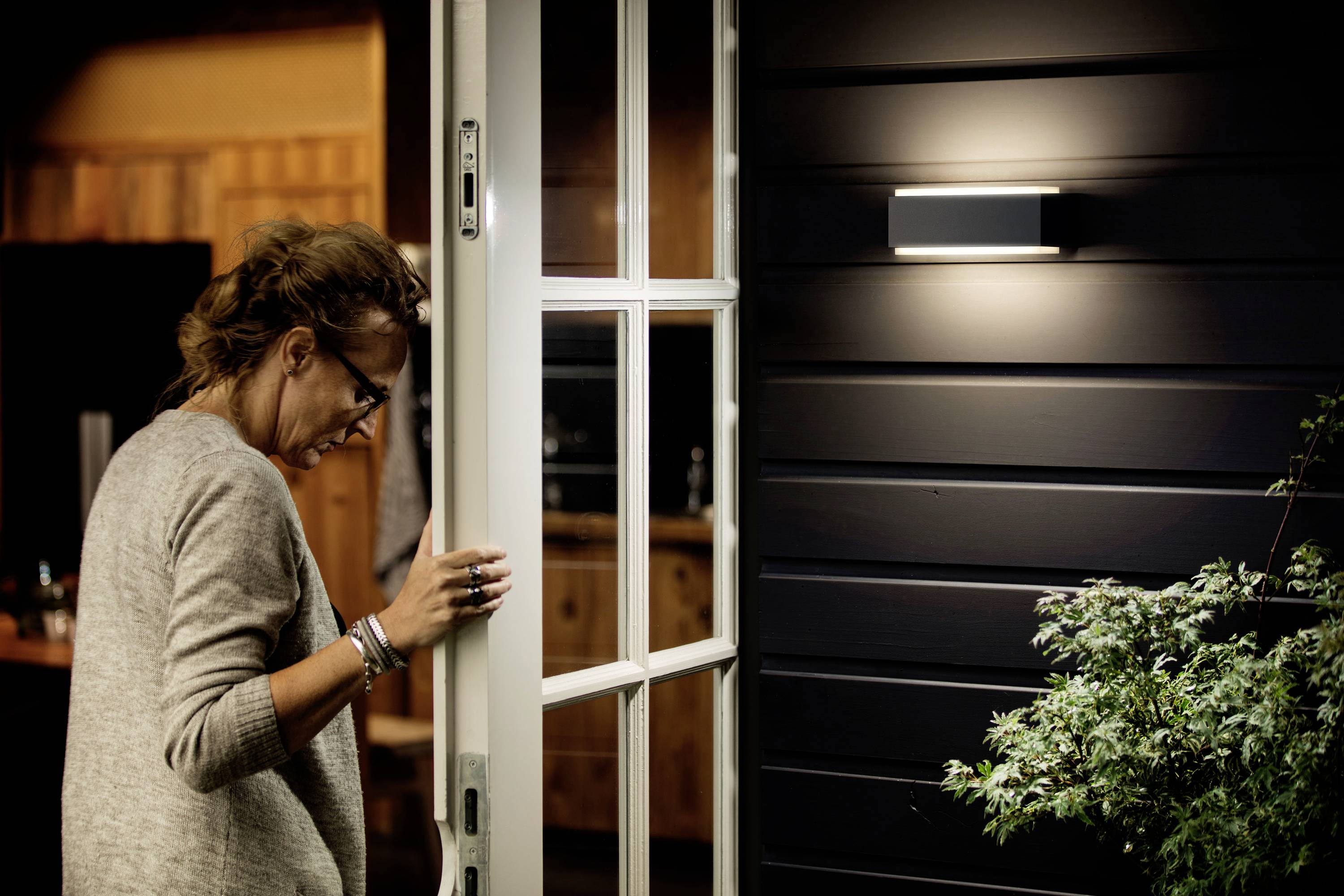 Eine Frau schließt eine weiße Glastür in einem beleuchteten Raum. Außen ist eine dunkle Wand mit einer Lampe, und eine Pflanze steht daneben.