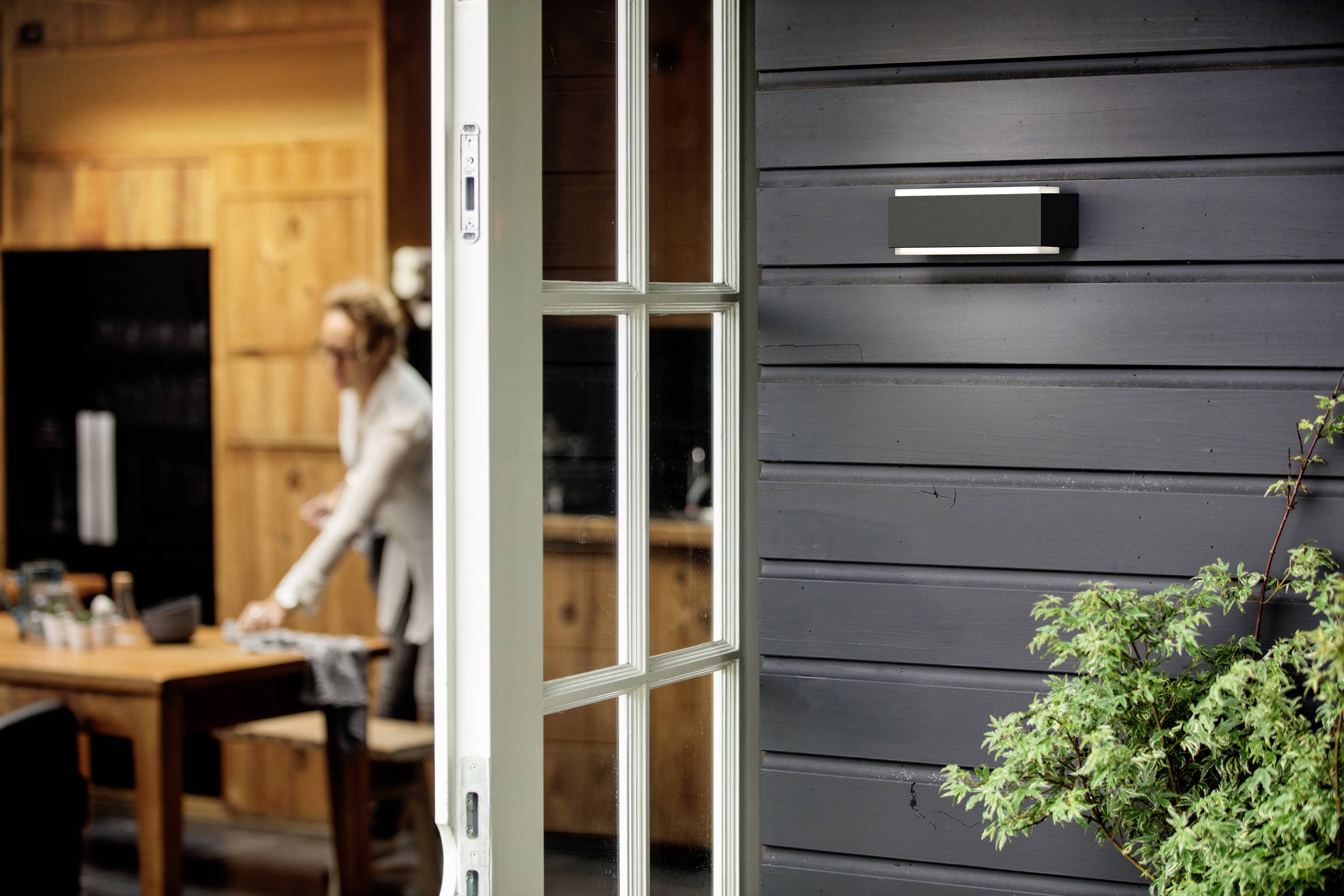 Ansicht einer geöffneten Glastür, die zu einem Raum mit einem Tisch im Hintergrund führt; ein Mensch ist im Raum unscharf sichtbar.