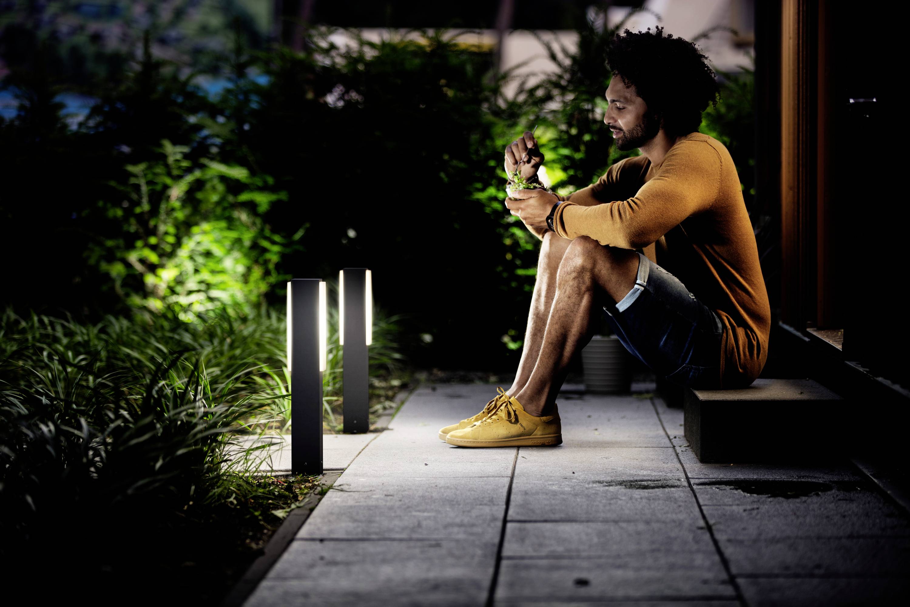 Ein Mann sitzt mit gekreuzten Beinen auf einer Terrasse neben beleuchteten Gartenleuchten und blickt auf ein Gerät in seiner Hand.